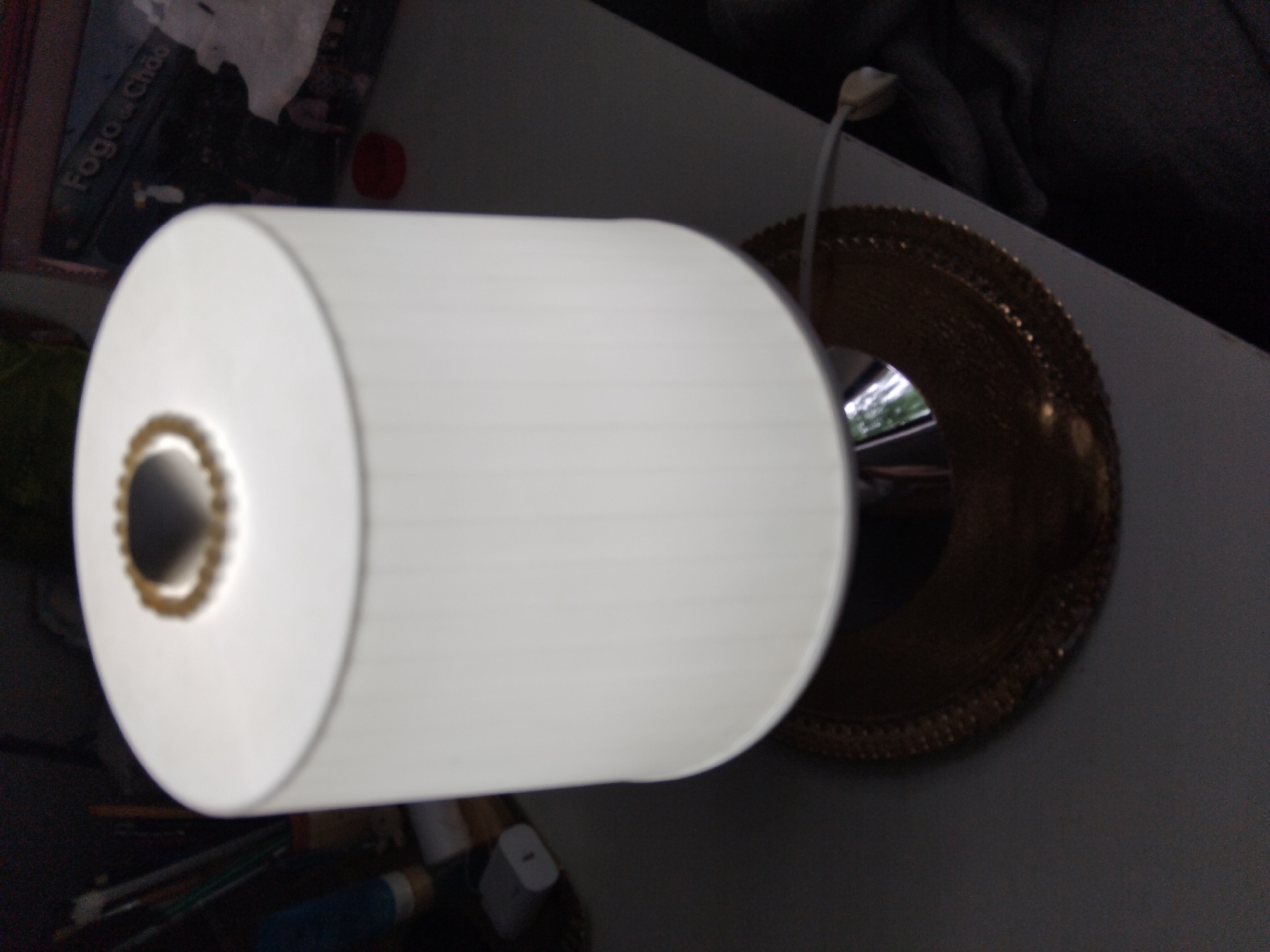 The image shows a bedside lamp with a white cylindrical shade and a gold base. The lamp is illuminated from within, casting a soft glow. On top of the lampshade, there is a decorative beaded band. The lamp is situated on a flat surface, likely a table or nightstand. In the background, out of focus, are various objects including what appears to be a framed picture or poster with Portuguese text that reads "Fogo de Chão", art supplies like pencils and brushes in a container, and a white electrical adapter. The overall impression is that of a domestic setting, possibly a bedroom. The image does not contain any specific landmarks or features that would allow for precise location identification within Vila Nova de Famalicão, Portugal.
