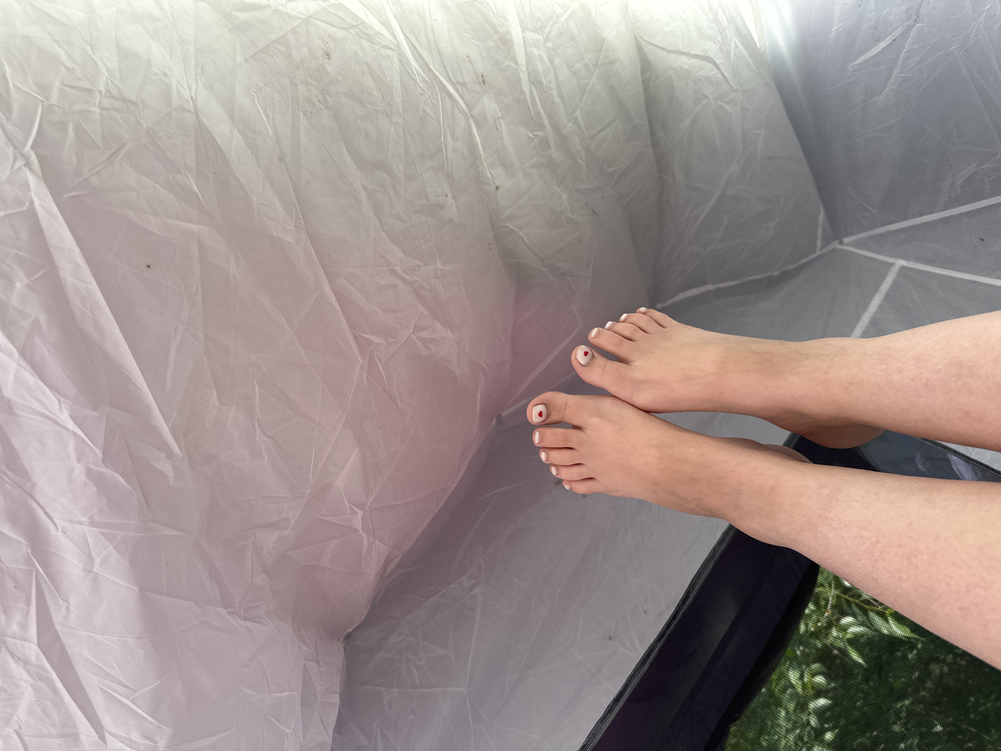 A high-angle shot shows two bare feet with white and red nail polish resting inside a tent. The tent's fabric is wrinkled and white. Green foliage is visible through the mesh window of the tent. The location is the city of Zhemchuzhny, Russia.