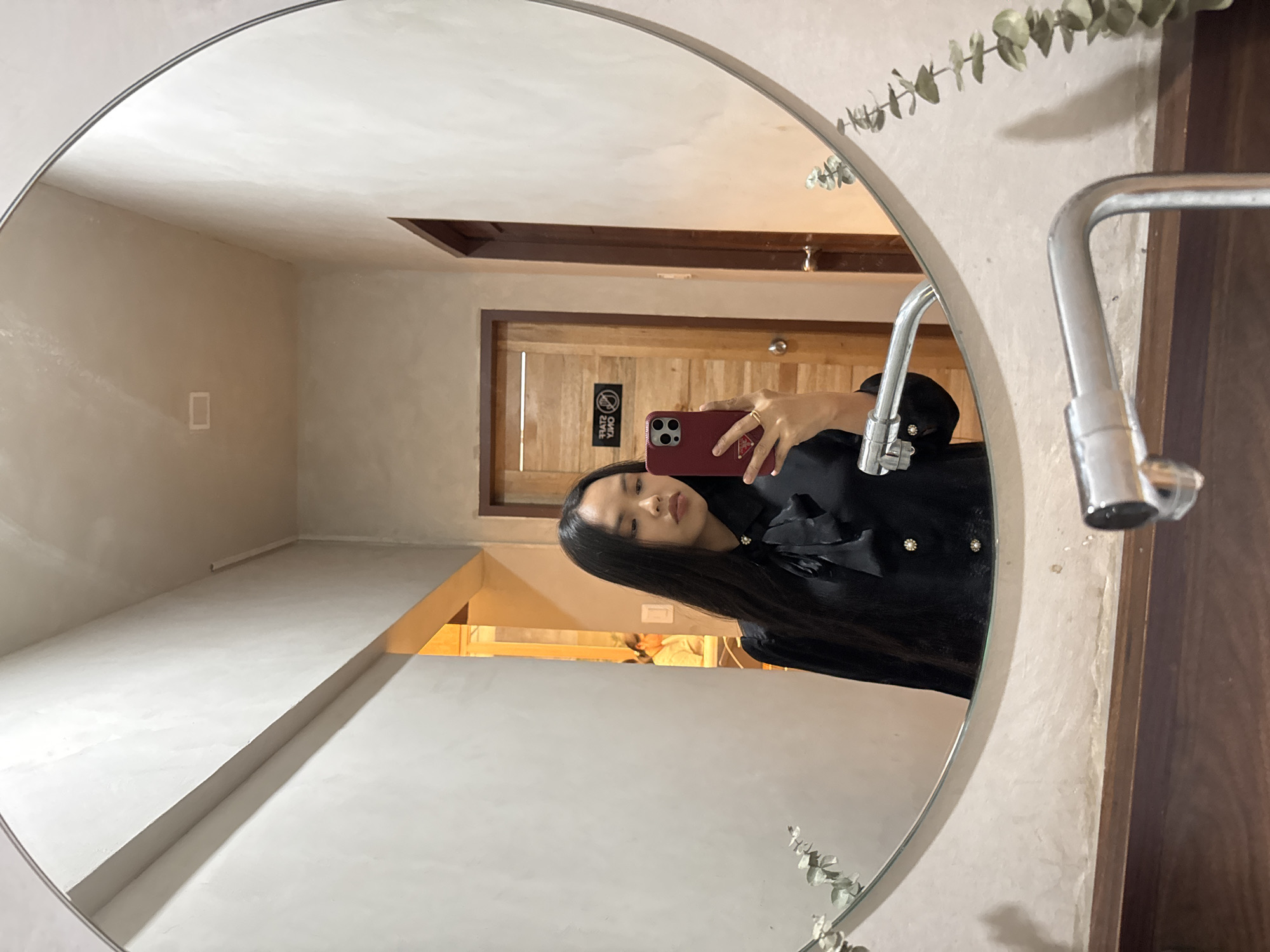 A woman is taking a selfie in a round mirror in a bathroom. The mirror is mounted on a wall with textured plaster. A silver faucet is visible on the right side of the mirror. The reflection shows a door in the background with a sign on it. The woman is holding a red phone with multiple camera lenses. She is wearing a black blouse with buttons and has long, dark hair. The lighting is warm. Based on additional information, this scene is located in Davao City, Philippines.
