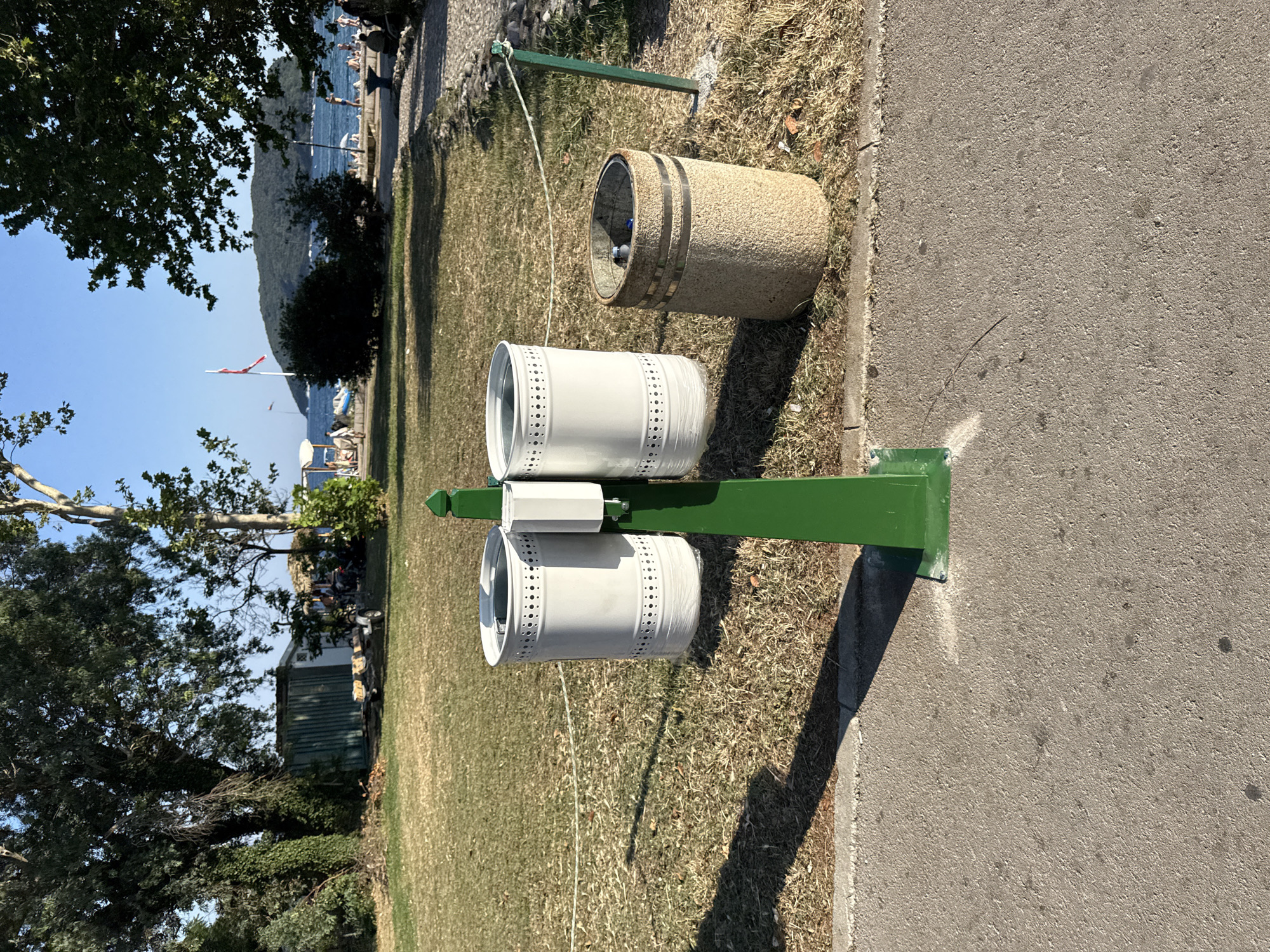 The image shows a green metal trash can holder with two bins. One bin is white and has a decorative pattern of small holes. The other bin, positioned above the white one, is made of a textured, sandy material with two metallic bands. Both bins appear to be empty. They are situated outdoors, adjacent to a paved walkway. Behind the trash cans, a grassy area extends towards a body of water, likely the sea, with a distant hill or mountain range visible. Sunlight casts shadows, suggesting it is daytime. In the background, there are trees, a flagpole with a flag, and some buildings. There are also some people visible near the water. The scene evokes a public outdoor space, possibly a park or promenade, in a sunny climate.
