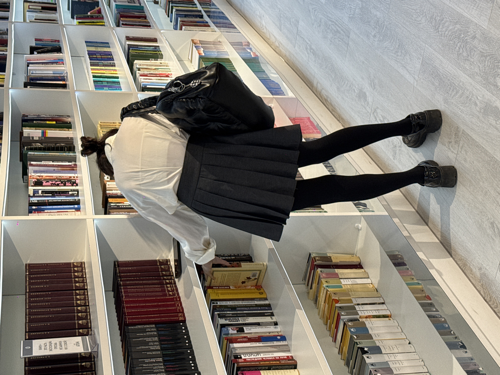 A young woman, dressed in a white collared shirt, a grey pleated skirt, black tights, and black platform shoes, is browsing a bookcase in a bookstore or library. She is wearing a large black handbag on her back. The shelves are full of books, many with Russian titles. The location is likely in Moscow, Russia, based on the language of the books. The woman appears to be looking at a book on the second to the bottom shelf, possibly to choose it. The floor is a light grey wood laminate.