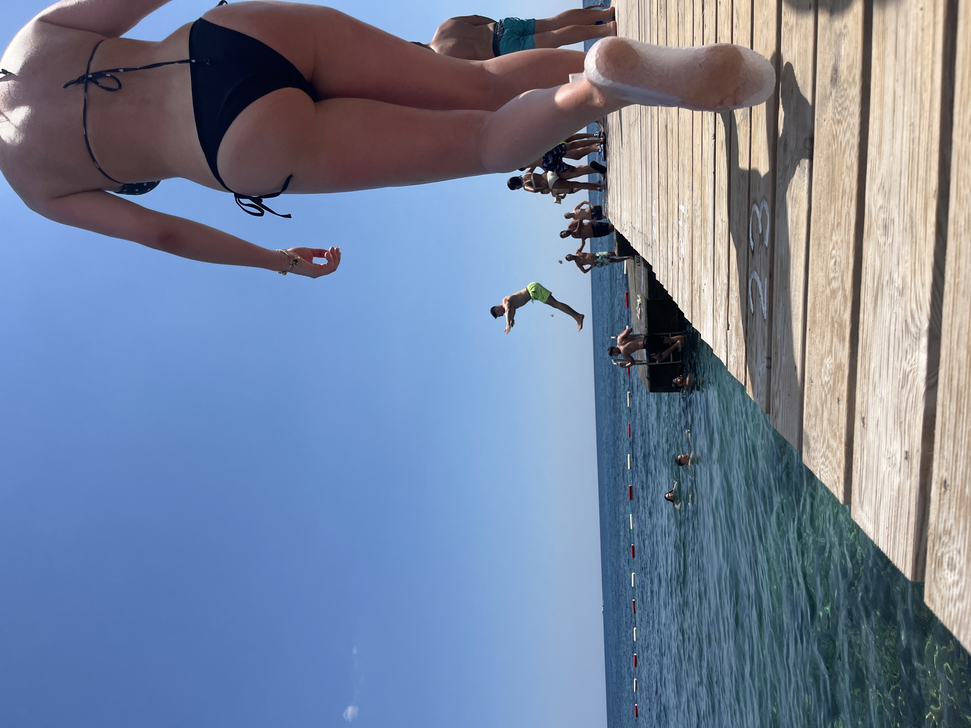 The image captures a sunny day at a popular swimming spot, likely on the coast of Budva, Montenegro, given the context provided. In the foreground, a person's lower legs and feet, clad in a black bikini bottom, are prominently displayed, walking away from the camera on a wooden pier. The pier extends out over clear, turquoise water. In the mid-ground, several other individuals are visible on and around the pier. One person is captured mid-air, jumping from the pier into the water, wearing lime green shorts. Others are standing on the pier, some appear to be watching the jumper, while others are preparing to enter the water. A few people are already in the water, swimming. A buoy line with red and white markers separates the swimming area from the open sea, which is calm and blue under a clear sky. The wooden pier has markings on it, including the numbers "23" painted on the planks. The overall scene depicts a lively and recreational atmosphere.