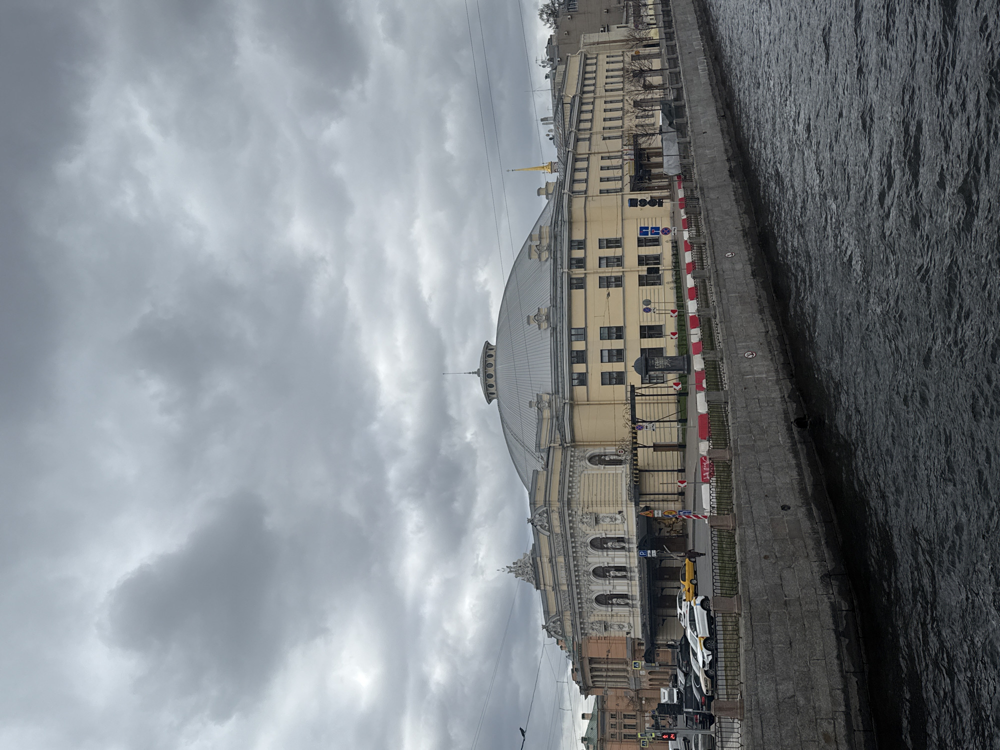 The image, taken sideways, captures an overcast urban scene in Saint Petersburg, Russia, alongside a body of water, likely a river or canal. The sky is dominated by thick, grey clouds, indicating a dull or rainy day.
A prominent, grand building with a large, light-colored facade and a distinctive domed roof occupies the left side of the frame. This ornate structure features classical architectural details, multiple windows, and decorative elements, strongly resembling the Bolshoi Saint Petersburg State Circus (Ciniselli Circus) on the Fontanka River. Behind it, a slender golden spire, possibly belonging to Mikhailovsky Castle, rises above the rooftops.
Separating the buildings from the dark, rippling water on the right is a wide, paved embankment or street. A stone wall with railings runs along the water's edge. Parked cars, including a yellow taxi and several white vehicles, line the street. Red and white striped barriers are visible along parts of the embankment. Various street signs are present, with a blue sign clearly displaying "ПАРКОВКА" (PARKOVKA - PARKING) alongside a white 'P' symbol. Another blue sign with a white arrow is also visible. The scene is static, depicting a typical cityscape on an overcast day, with no specific human activity taking precedence.