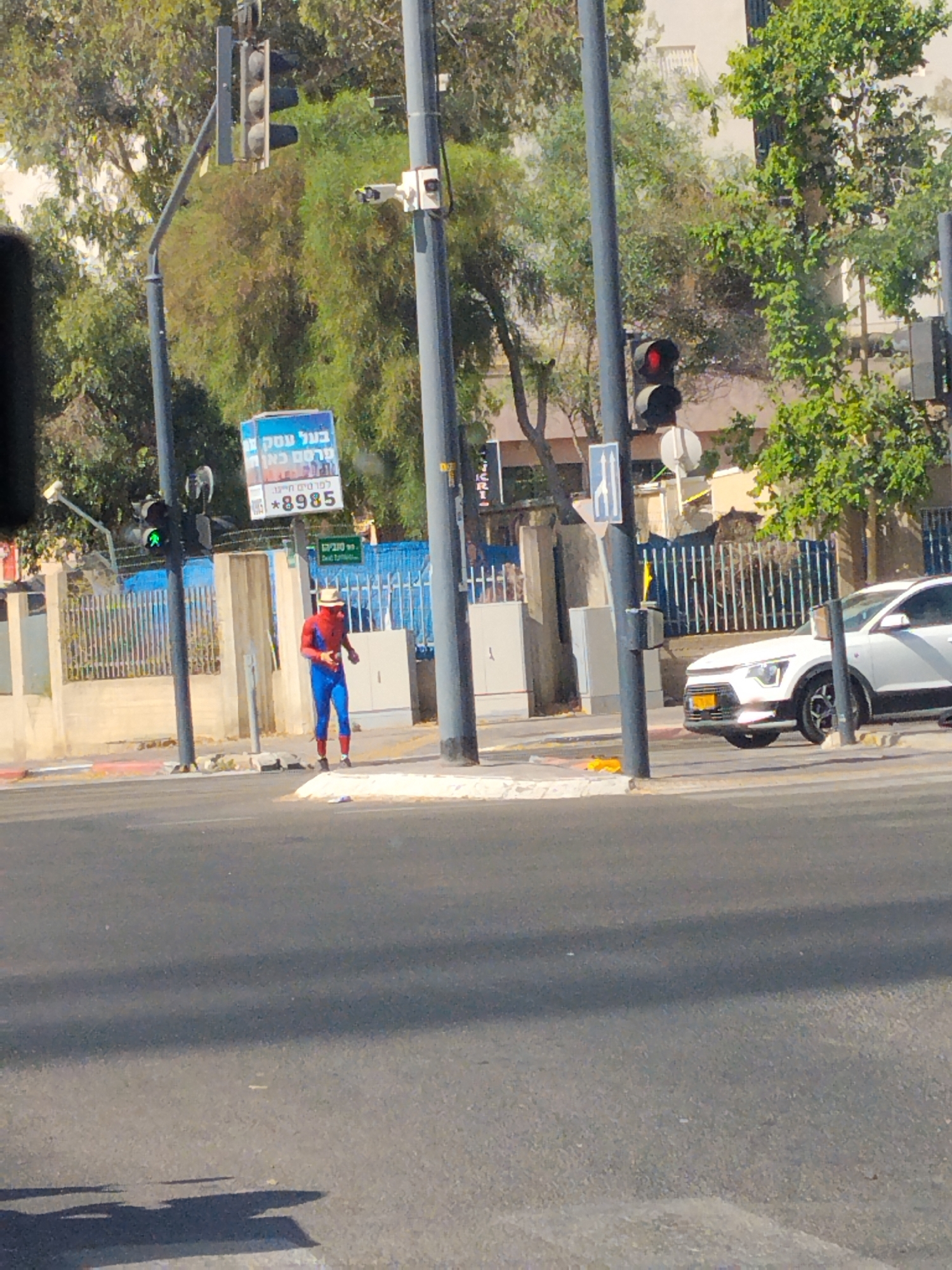 A person dressed as Spider-Man stands on a pedestrian crossing at an intersection in Be'er-Sheva, Israel. The person is wearing a red and blue Spider-Man costume, red shoes, and a straw hat. They are holding what appears to be a phone or remote in their right hand. To the left of the person, a traffic light shows a green signal. Behind the person, there is a blue fence and buildings. In the background, trees and streetlights are visible. A white car is stopped at the intersection to the right of the Spider-Man figure. A billboard displaying Hebrew text and a phone number is mounted on a pole near the traffic light. The ground is paved and there are shadows cast by the sun. The scene suggests a sunny day in a city environment.