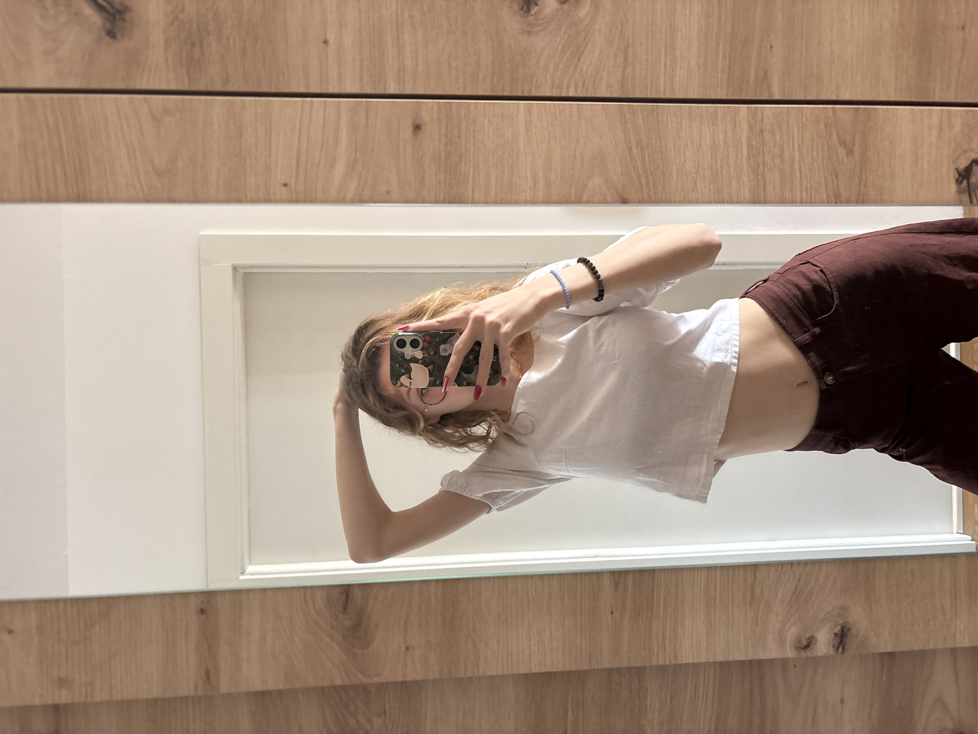 A young person is taking a mirror selfie. They are wearing a white, short-sleeved crop top and maroon pants. The phone is held up, obscuring the person's face. Two bracelets are visible on their wrist. The mirror has a white frame and is set against a wood-paneled wall. There is no information available to indicate the location is Kragujevac, Serbia.