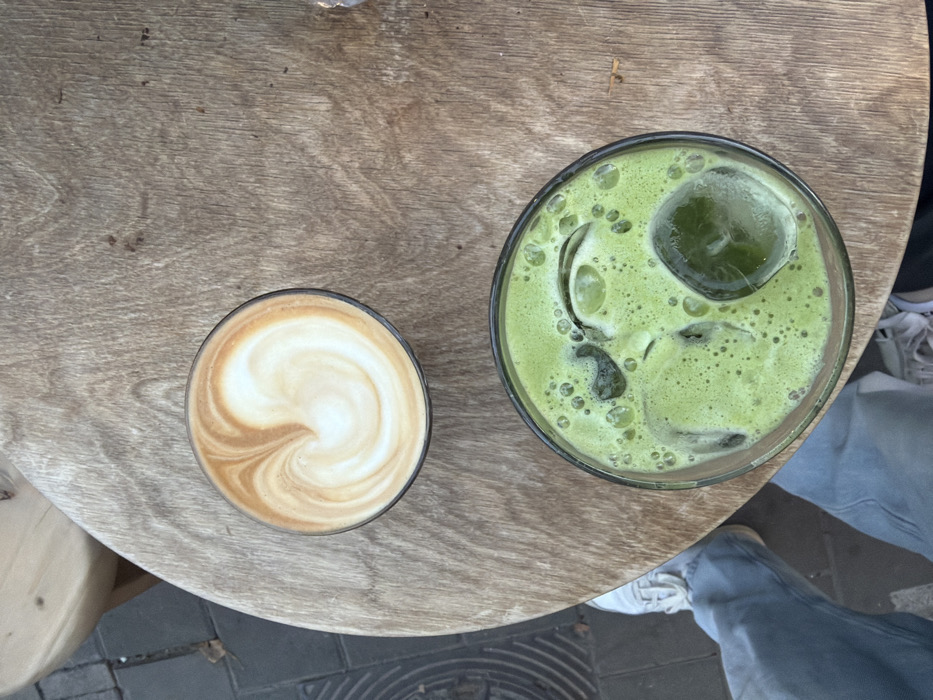 An overhead view captures two beverages on a round, light-colored wooden table in Tel-Aviv, Israel. The table surface displays visible wood grain and minor imperfections. In the upper portion, a clear glass contains a coffee beverage topped with white foam and a brown heart-shaped latte art design. Below it, a second clear glass holds a vibrant green, iced liquid with several ice cubes, one of which is prominently larger and spherical. To the bottom left of the frame, the lower torso and legs of an individual are partially visible, clad in light blue denim jeans and white athletic shoes, positioned beneath the table. The ground adjacent to the table is paved with dark grey material, and a segment of a circular metal grate is observable on the extreme left.