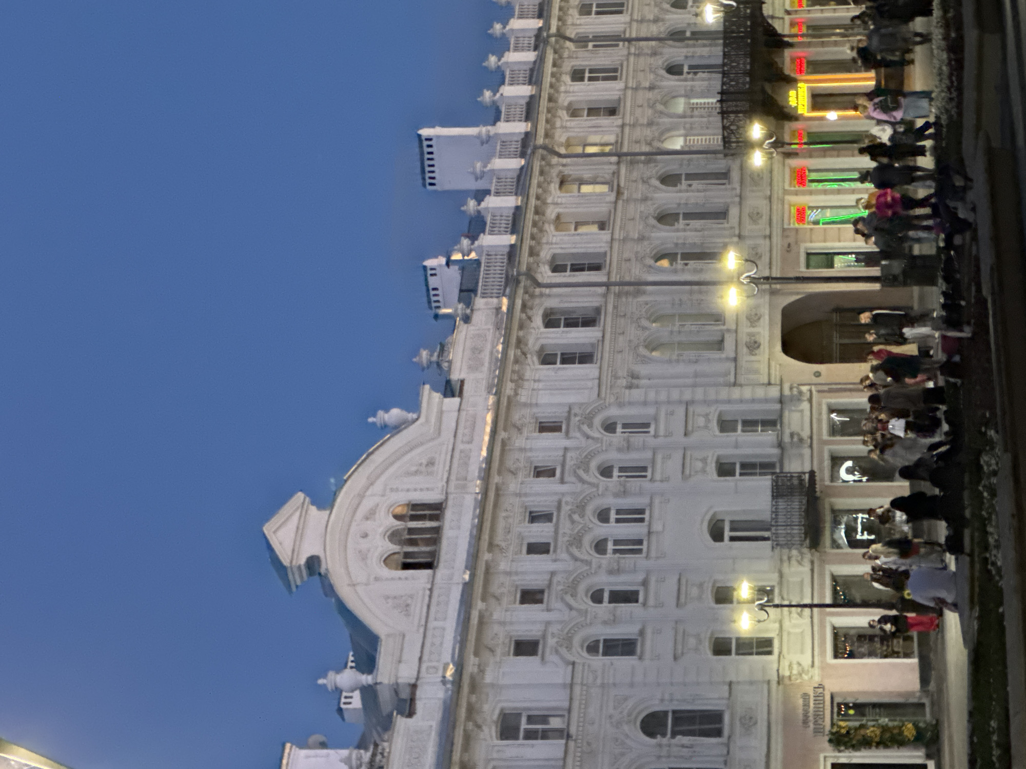 The image depicts a twilight scene of a busy street in Nizhny Novgorod, Russia. In the foreground, a group of people are gathered on a paved area, some sitting while others stand. The background is dominated by a grand, ornate white building with multiple stories, featuring numerous windows and intricate architectural details. Streetlights illuminate the scene, casting a warm glow. Several shops are visible at the ground level of the building, with some displaying colorful neon signs. The sky above is a deep blue, indicating the transition from day to night. The overall impression is one of urban activity and historical architecture.