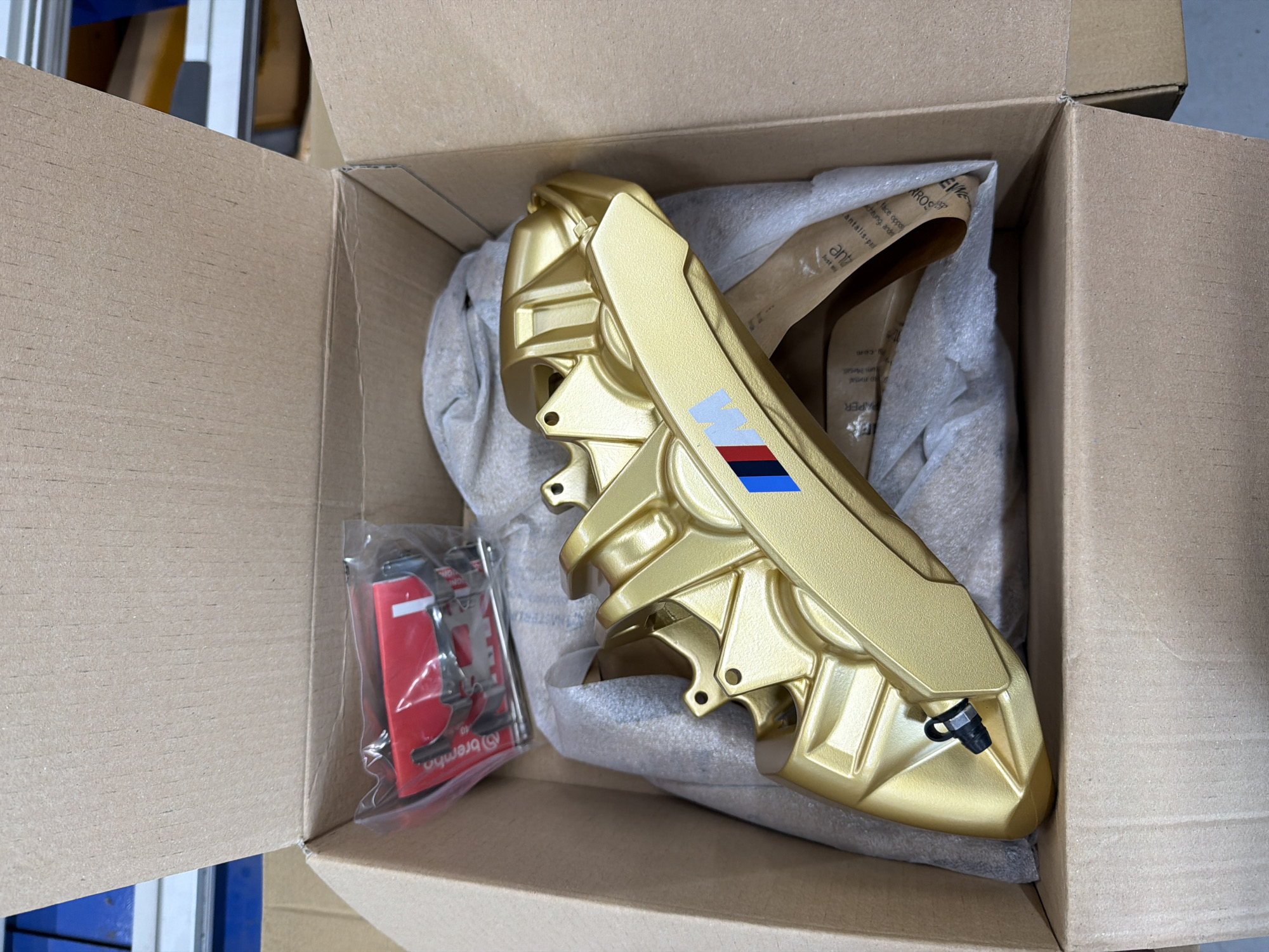 An overhead shot captures a brand new golden car brake caliper resting inside a plain brown cardboard box. The caliper, prominently featured, is a striking metallic gold/bronze color and bears the iconic BMW "M" logo with light blue, dark blue, and red stripes on its side. It is cushioned by grey protective packaging material, likely bubble wrap or foam, and crumpled brown packing paper.
To the left of the caliper, a small clear plastic bag contains additional brake hardware, including red and silver components. Visible text on one of these red parts confirms the brand "brembo." The box itself appears fresh and undamaged, suggesting the caliper has just been unboxed or is awaiting installation.
Visible text on the crumpled packing paper includes partial words and phrases such as "com", "EV", "AROS", "face oppos", "chitung, ande", "antalis-pal", "ant", "Jost", and "PAPER". The background outside the box is out of focus, showing hints of blue and white structures, suggesting an indoor setting like a workshop or garage. The lighting is bright and even, highlighting the new condition of the car part.