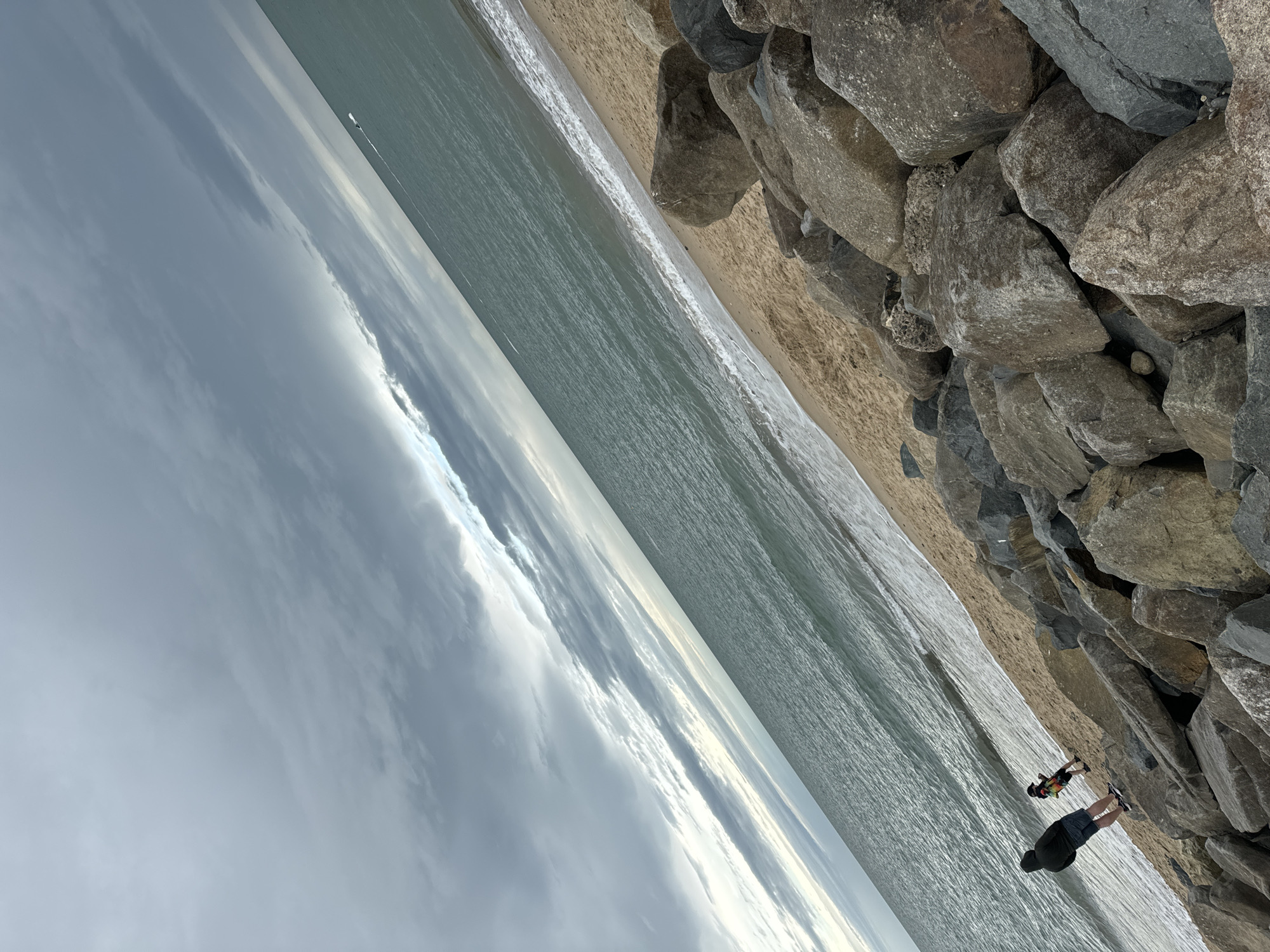 The image depicts a view of the sea from a rocky shoreline in Courtown, Ireland. Two individuals are visible: an adult wearing a dark hooded jacket and shorts, standing on the rocks, and a smaller figure, possibly a child, in colorful clothing, walking on the sandy beach. The sea is calm with gentle waves breaking on the shore. The sky is overcast with dark clouds, suggesting potential rain or a stormy weather front. The foreground is dominated by large, grey rocks forming a breakwater, with a sandy beach stretching alongside it. The overall scene conveys a sense of coastal natural beauty under a dramatic sky.