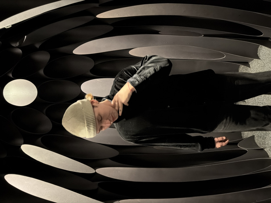 The image displays a young adult male standing within a distinctive architectural space at the Sense Hotel in Sofia, Bulgaria. The subject wears a light beige ribbed beanie, a dark, long-sleeved button-up shirt with a soft, possibly velvet texture, and dark trousers. A thin red string is visible on his left wrist, and an earring is present in his left ear. His head is angled downwards and slightly to the right, with his gaze directed towards the lower right. His left hand is raised, with fingers bent, creating a 'V' shape with his thumb and index finger, and touching his neck/jawline. The background consists of a dark grey wall featuring a repeating pattern of recessed, elongated oval shapes. Several of these shapes contain integrated light sources, with two prominent bright white circular lights visible towards the top. This geometric pattern creates an illusion of depth and a slightly curved, tunnel-like environment. The floor is a light grey, textured, speckled surface. The scene is illuminated by the integrated lights, casting soft light and shadows.