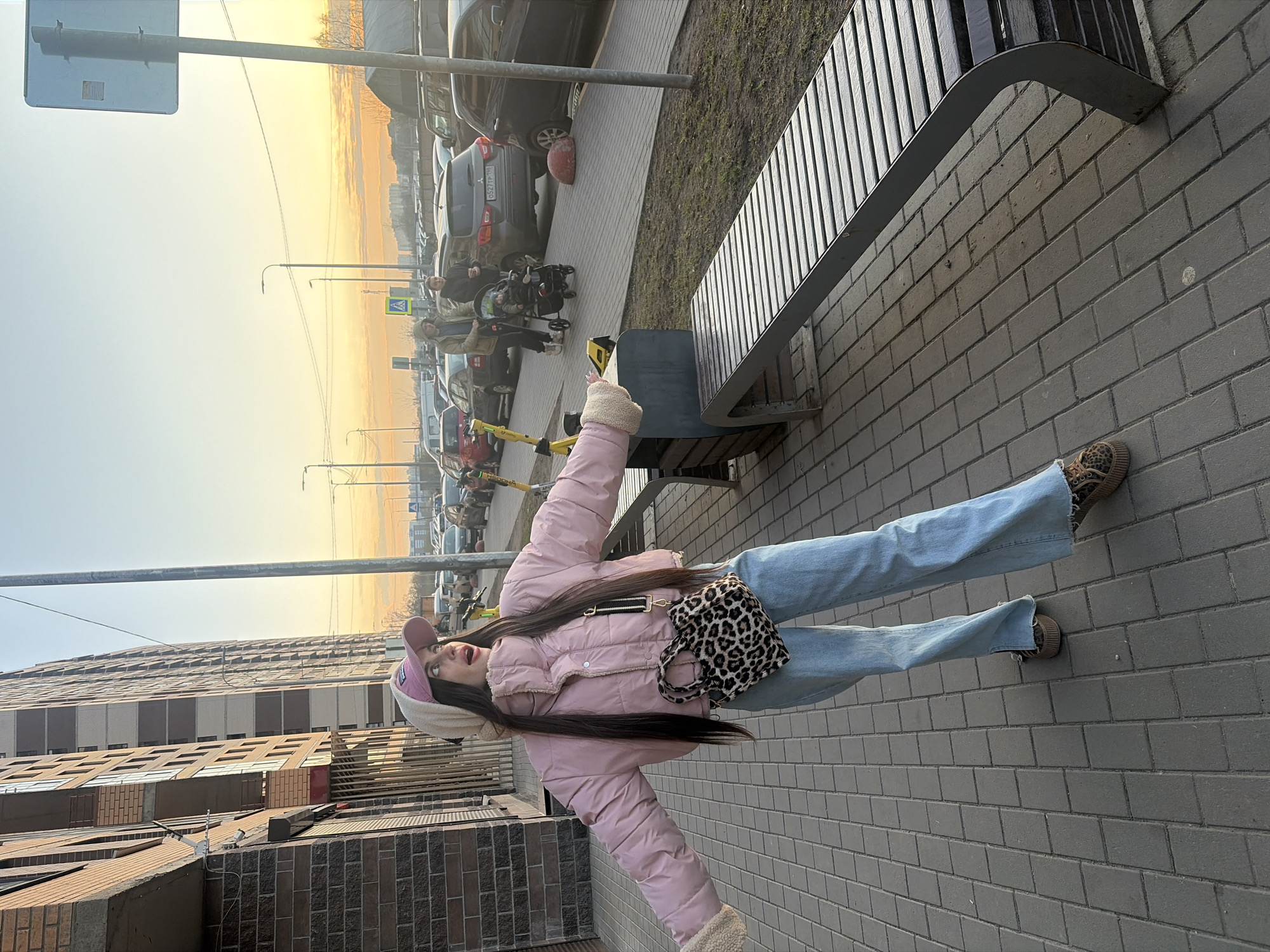 The image captures a woman standing on a sidewalk in what appears to be a city street, likely in Saint Petersburg, Russia. The scene is outdoors, with a clear sky visible in the background. The woman is dressed in a pink puffer jacket, light blue jeans, and leopard print sneakers. She is also wearing a pink and white cap and carrying a leopard print bag.
In the background, there are parked cars and a building. The setting sun adds a warm glow to the overall scene. Additionally, there is a bench next to the woman, along with some street furniture and infrastructure, such as poles and signs.