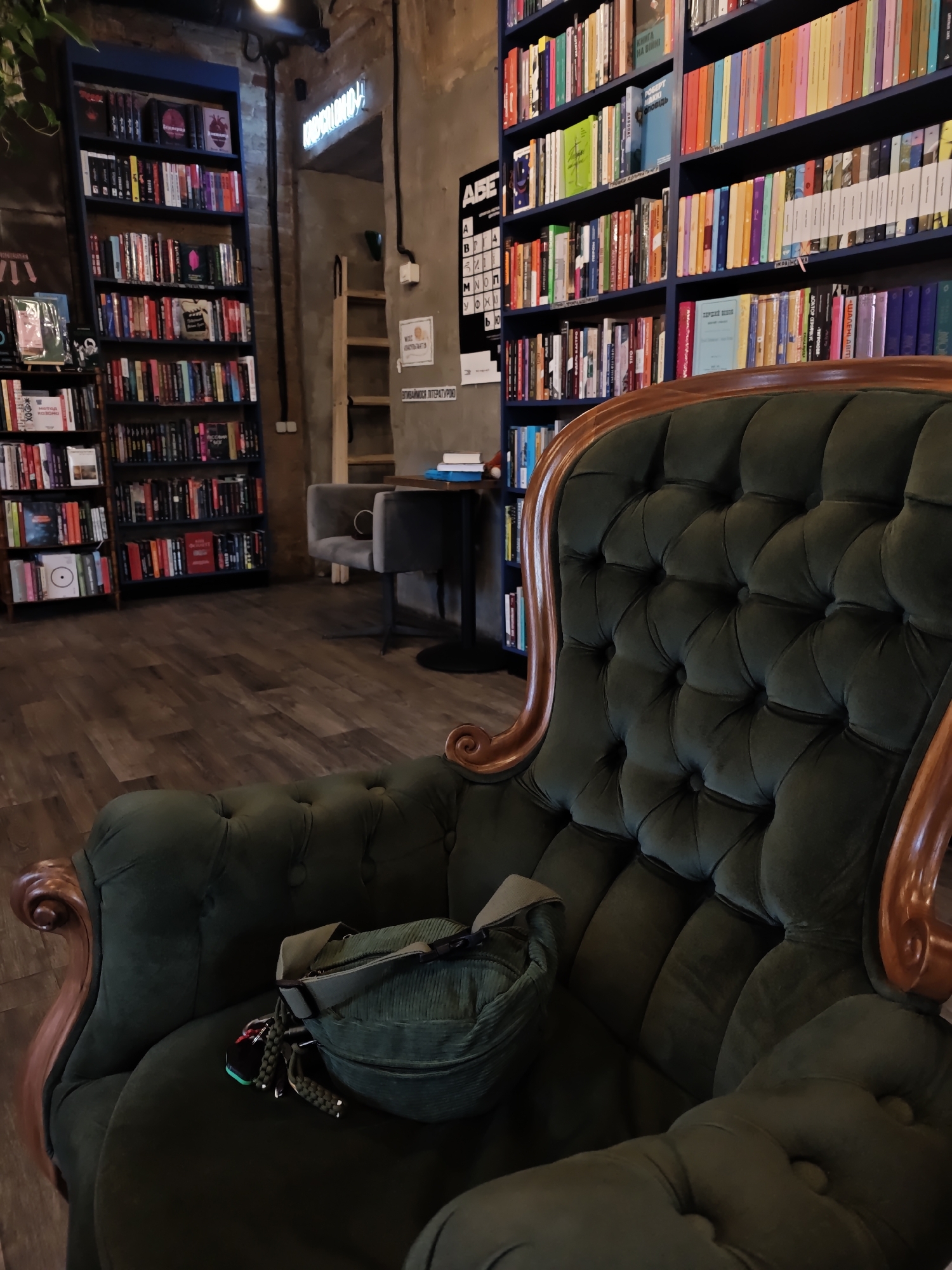 The image captures an indoor scene, likely a bookstore or cafe, in Kyiv, Ukraine. In the foreground, a plush, dark green tufted armchair with ornate wooden trim is prominently featured. A green corduroy fanny pack rests on the seat of the armchair.
In the background, tall bookshelves filled with books are visible on both the left and right sides of the frame. The shelves are organized with books of various colors and sizes. To the left, a blue bookshelf unit stands against a wall. To the right, a wider bookshelf unit displays a multitude of colorful books.
Between the bookshelves, a ladder-like shelf unit is positioned. Further back, a small grey chair is placed near a table with two books on it. The walls appear to be made of brick or concrete. A neon sign with Cyrillic lettering is visible on the wall above the ladder shelf. The floor is covered with wooden planks. The overall ambiance suggests a cozy and literary setting.