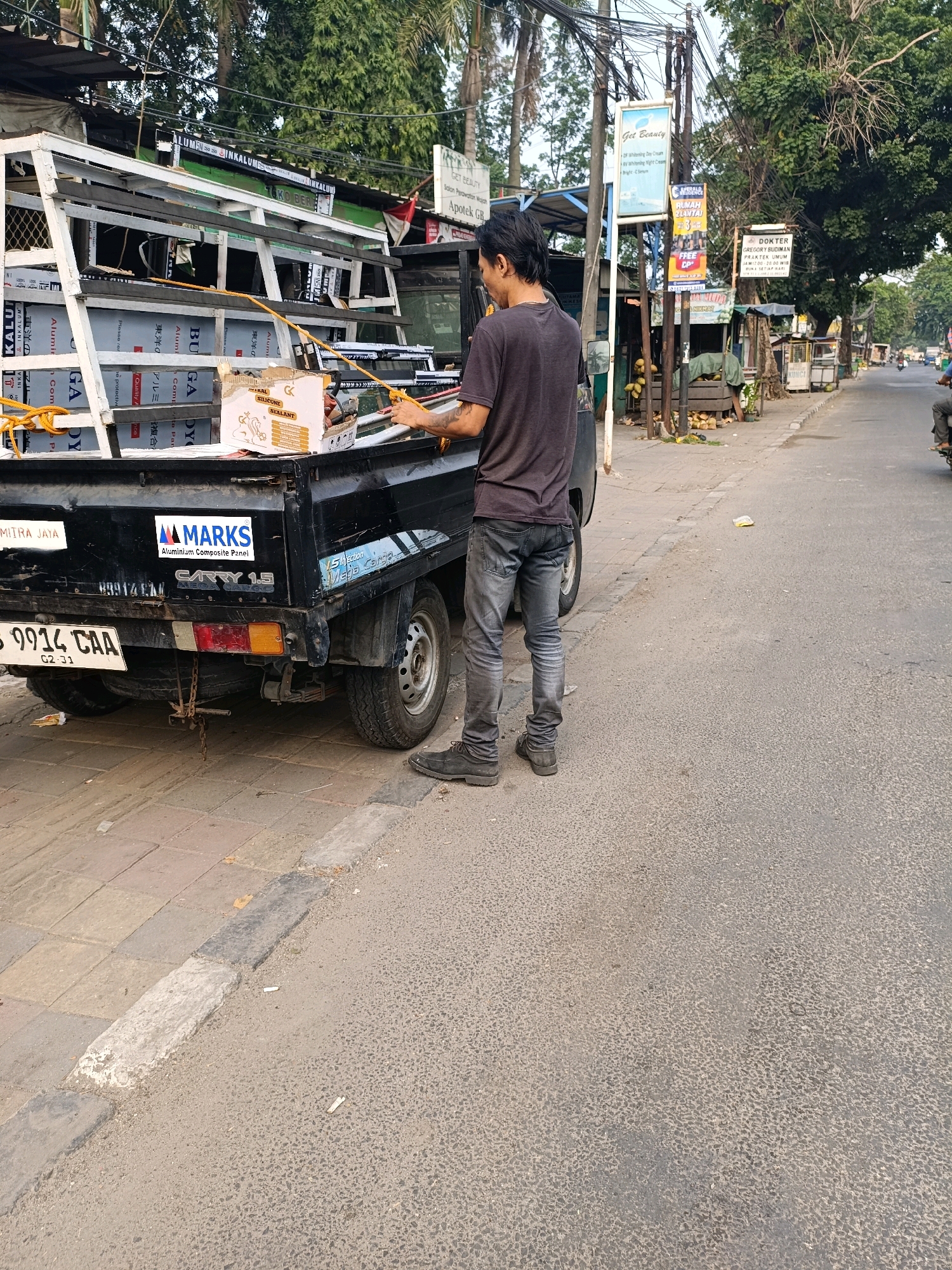 A young man, wearing a dark grey t-shirt and faded grey jeans, stands on a paved road next to the back of a black pickup truck. The truck is loaded with aluminum composite panels, stacked on a metal frame. The man is holding a yellow rope and appears to be securing or adjusting the cargo. The license plate of the truck reads "B 9914 CAA". The scene takes place on a street in Tangerang, Indonesia, with trees and various roadside businesses visible in the background. The road is asphalt, and there is a sidewalk with paving stones. The overall atmosphere suggests a typical street scene in an Indonesian city, possibly during the day, with moderate traffic implied by the presence of other vehicles in the distance.