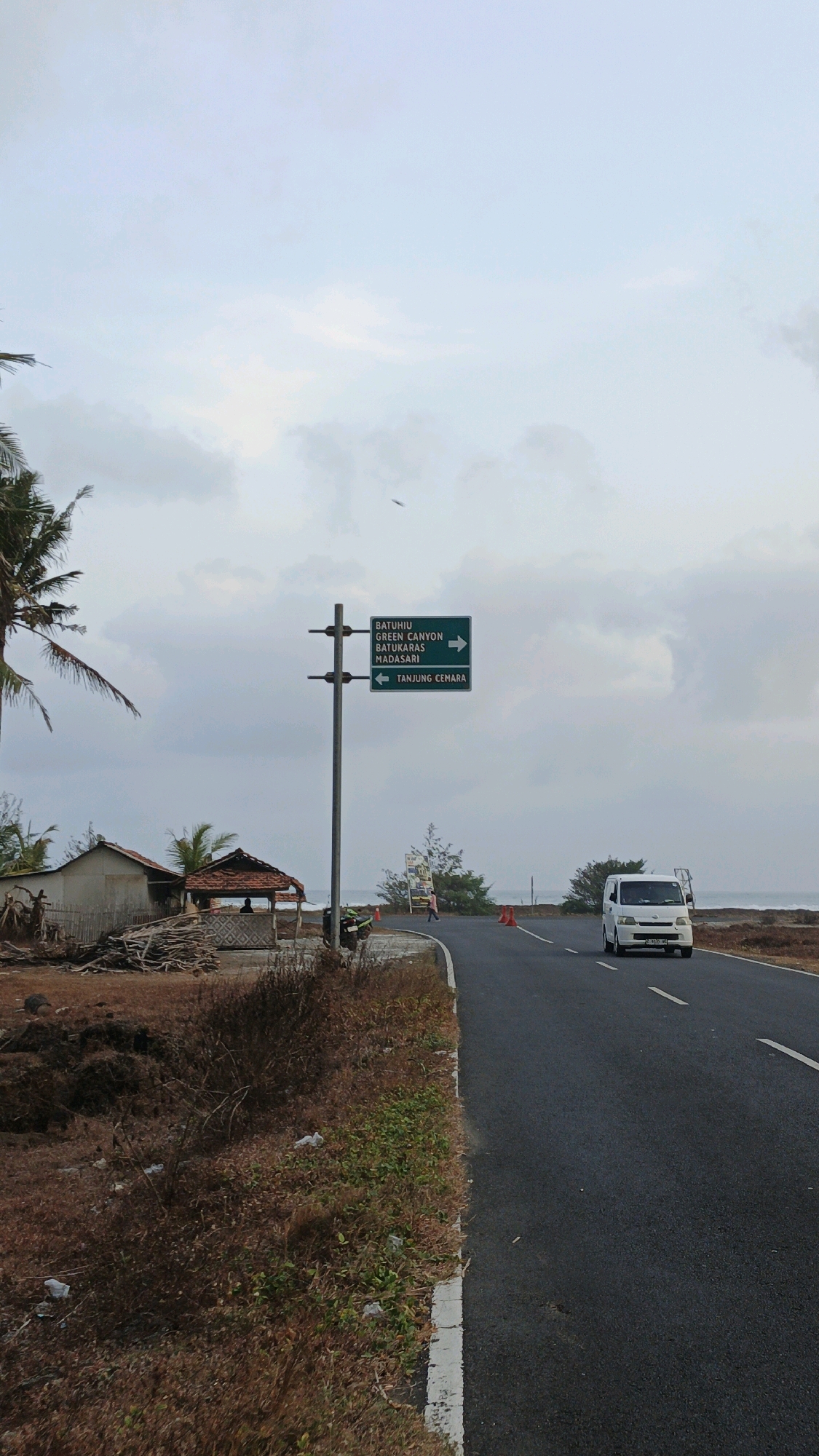 In Sukaresik, Indonesia, a roadside scene unfolds under a cloudy sky. A paved road curves gently to the right, with a white van traveling away from the viewer. To the left of the road, a signpost stands with two green directional signs. The top sign lists "BATUHIU," "GREEN CANYON," "BATUKARAS," and "MADASARI," with an arrow pointing to the right. The bottom sign reads "TANJUNG CEMARA" with an arrow pointing to the left. In the foreground, to the left of the road, are two small, rustic structures and a pile of wood. The ground is covered in dry grass and some scattered white plastic debris. Palm trees are visible on the far left. Beyond the road, the ocean stretches to the horizon. Orange traffic cones are placed along the edge of the road. A person walks in the distance near the cones.
