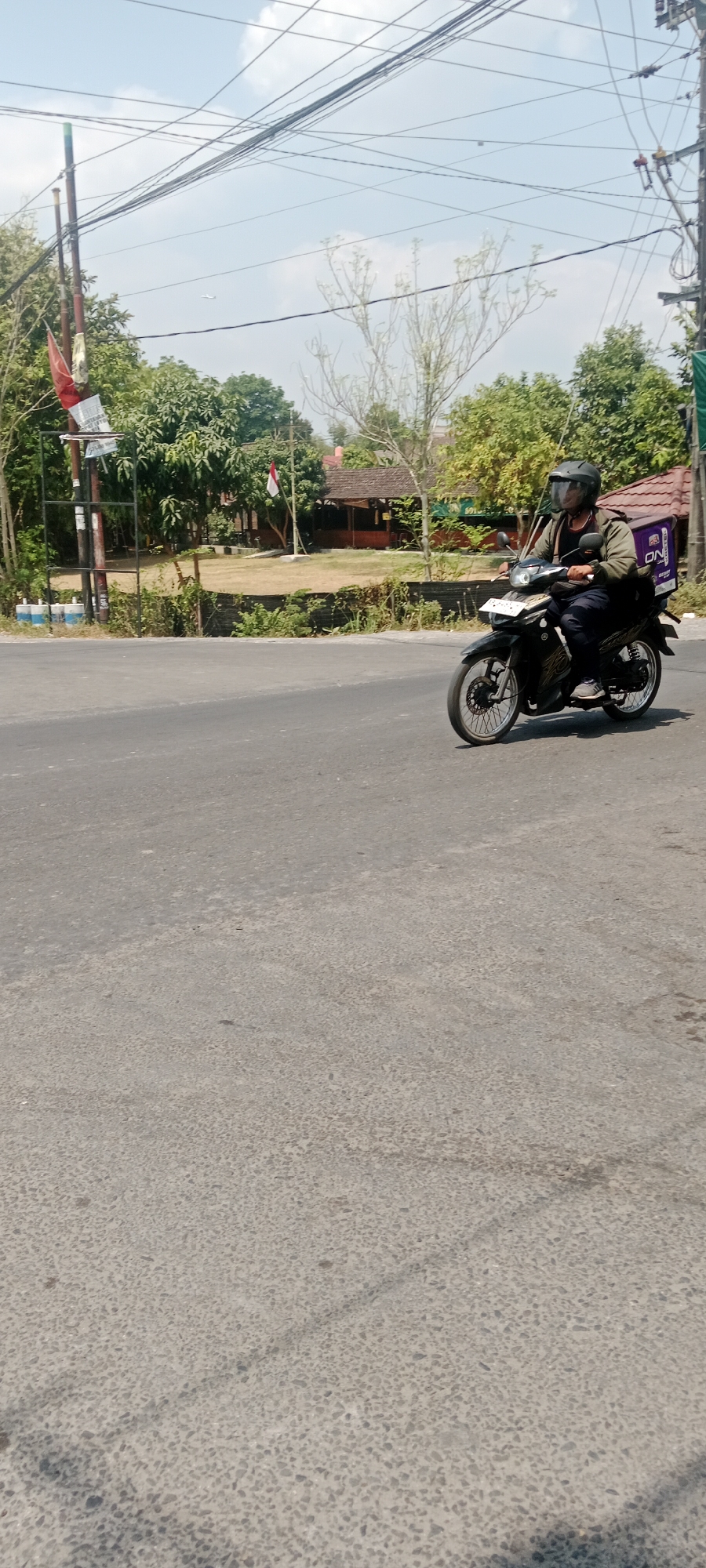 A delivery person wearing a helmet and a dark jacket rides a dark-colored motorcycle on a paved road. The motorcycle has a purple cargo box with the "ON" logo on the back. The delivery person is looking forward as they ride. In the background, there are trees, a building with a tiled roof, and utility poles with many wires. A flag, likely the Indonesian flag, is visible on a pole. The sky is clear and blue, suggesting it is a sunny day. The scene appears to be in an urban or suburban area of Maguwoharjo, Indonesia, possibly near a residential area or commercial establishment.