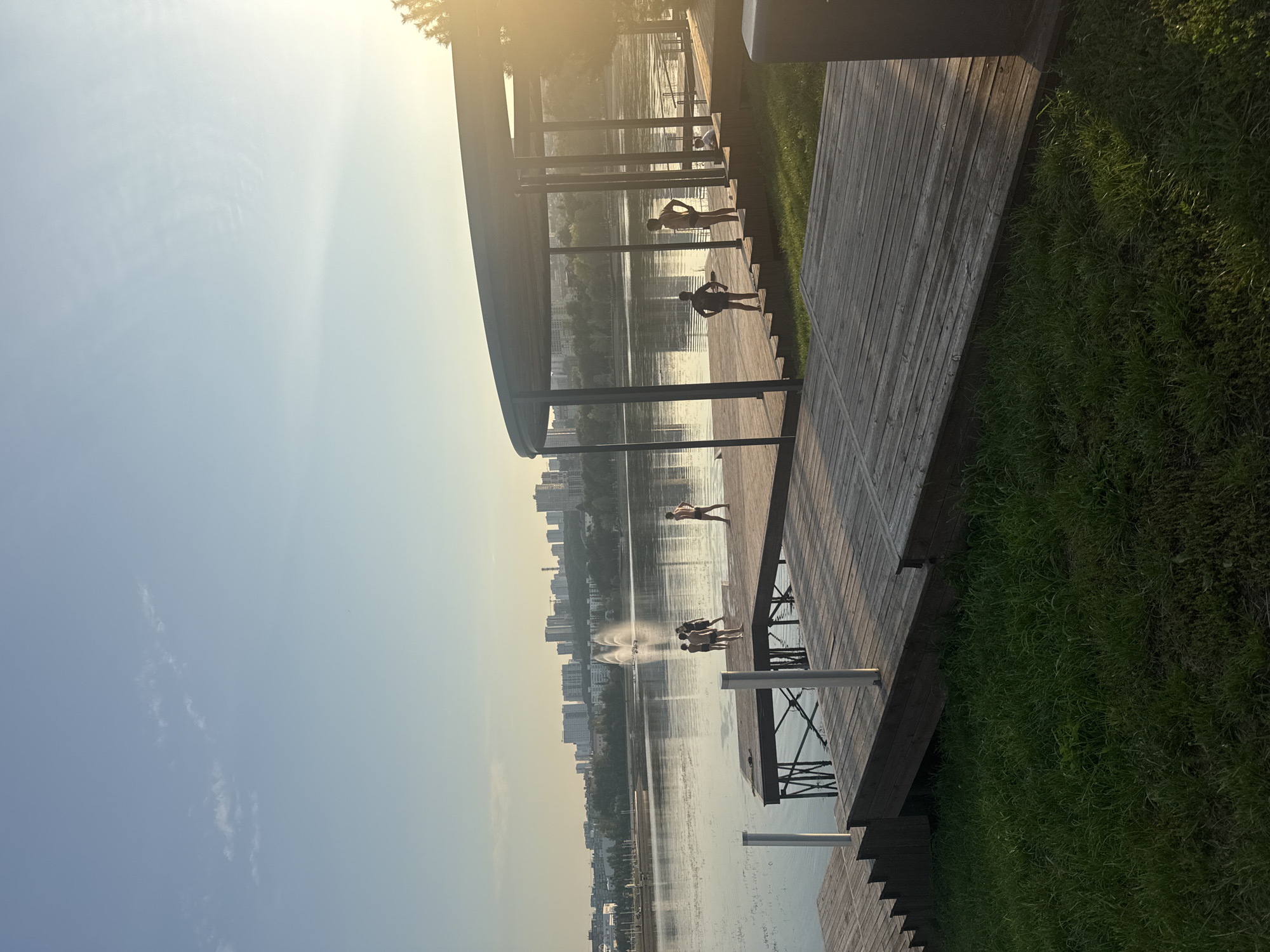 The image shows a waterfront area in the city of Ufa, Russia. In the foreground, there is a wooden deck with a circular canopy structure. Several people, appearing to be children, are visible on the deck and a wooden pier extending into the water. Some are standing, while others are gathered in small groups. In the background, across a body of water, a cityscape with numerous buildings and a fountain is visible. The sky is clear with some clouds, and the sun is setting, casting a warm light on the scene. The overall setting suggests a public recreational area by the water, possibly a park or beach.