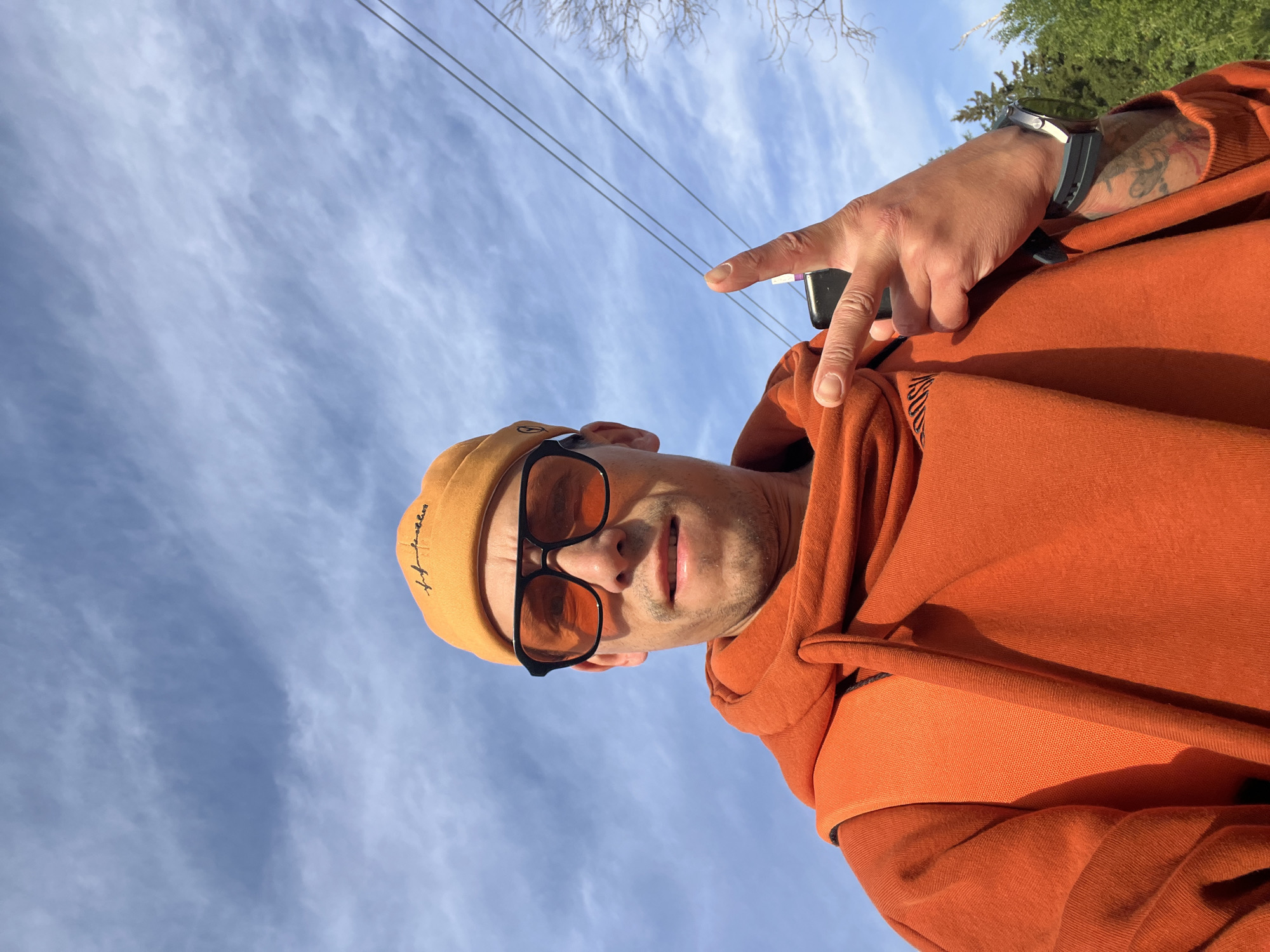 The image is a selfie of a man, taken outdoors in Chaikovsky, Russia, from a low-angle perspective looking upwards. The man is centered in the frame against a bright, mostly blue sky with scattered wispy clouds.
He is wearing an orange hooded sweatshirt and an orange beanie, which has dark script text on its front, appearing to read "Hustle Hard." He is also wearing black-framed sunglasses with orange-tinted lenses. His face shows a light beard or stubble, and his mouth is slightly open, suggesting a relaxed or subtly smiling expression.
The man's left arm is visible, with a smart watch on his wrist and a tattoo on his forearm. His right hand is raised towards the camera, forming a peace sign (V-sign) with his index and middle fingers. He appears to be holding a small, dark electronic device between his fingers.
In the background, beyond the man, the sky dominates the scene. Some bare tree branches are visible on the left side, indicating possibly late winter or early spring, along with hints of green foliage from evergreen trees on the right. A few thin utility or power lines stretch across the upper part of the sky. The bright daylight suggests the photograph was taken during the late morning or afternoon on a clear day.