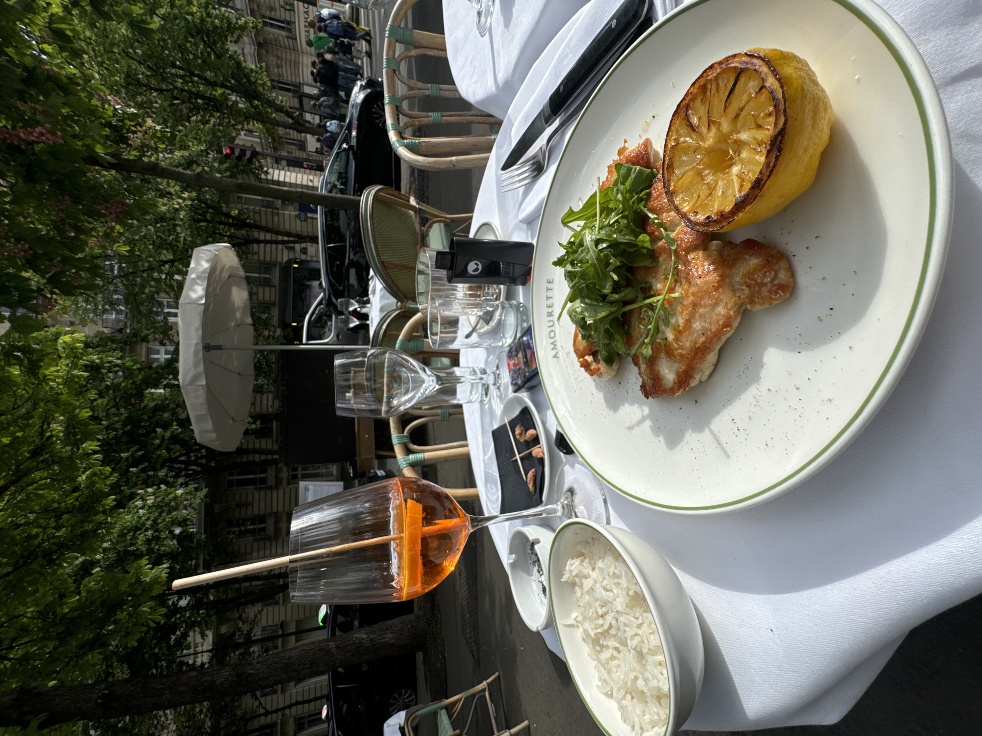 This image captures a vibrant outdoor dining scene at a café in Paris, France. The perspective is from a diner's eye level, looking across a table set for a meal.
In the foreground, a white tablecloth covers a table where various dishes and drinks are placed. The main subject is a white plate featuring a serving of grilled chicken or fish, topped with fresh arugula and accompanied by a charred lemon half. The word "AMOURETTE" is visible in green text along the rim of this plate. To the right, a small white bowl contains white rice. On the left, a tall glass holds an orange-hued beverage, likely an Aperol Spritz, with an orange slice and a straw, suggesting a refreshing drink for a sunny day. Other empty glasses, a fork, and a knife are also present on the table, along with a small black object, possibly a portable charger. Another small, dark plate holds what appear to be nuts or appetizers.
The setting is an outdoor area, indicated by the surrounding greenery and street elements. Behind the table, classic Parisian wicker chairs are arranged, and a white, folded umbrella can be seen. Large, leafy trees with some pink blossoms line the street, partially obscuring the background buildings. These buildings display typical Parisian architectural styles. Further in the background, blurry figures of people can be seen walking on the sidewalk, and cars are parked or passing by. One building in the background has partially visible text "MARPAC".
The scene depicts a leisurely meal or drink on a bright, sunny day, suggested by the strong light and distinct shadows. The overall atmosphere is relaxed and enjoyable, characteristic of a European café experience. The image evokes a pleasant outdoor moment in a bustling city environment.
