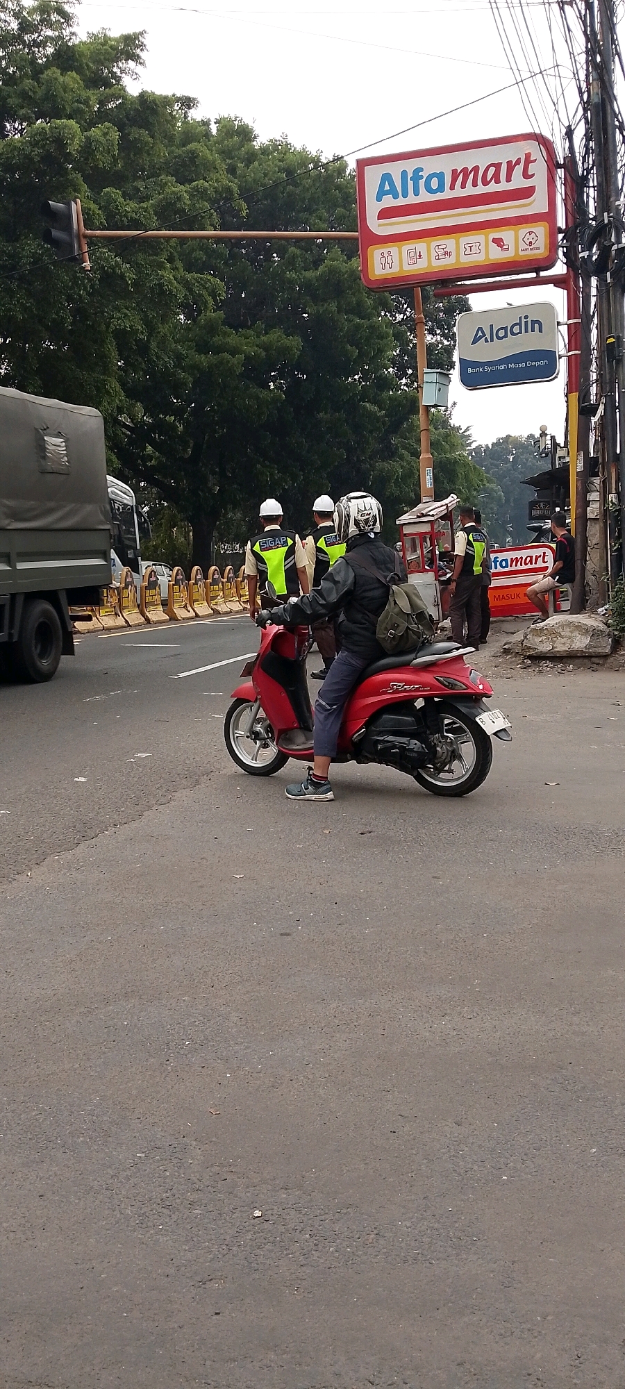 In Depok, Indonesia, a street scene unfolds with a red scooter in the foreground, ridden by an individual wearing a dark jacket, blue pants, and a white helmet with a black graphic. The rider is positioned on the right side of the road, facing left. Behind the scooter, four individuals in fluorescent yellow vests and white helmets stand near a small food stall. Two of them are wearing vests with "SIGAP" printed on them. A large grey truck is visible to the far left of the frame. Above the scene, an Alfamart sign is prominently displayed, accompanied by an "Aladin" sign below it, which indicates "Bank Syariah Masa Depan." The environment appears to be a busy road with trees lining the background. A traffic light is visible on the left side, and power lines crisscross the sky. Yellow barricades are positioned on the side of the road.