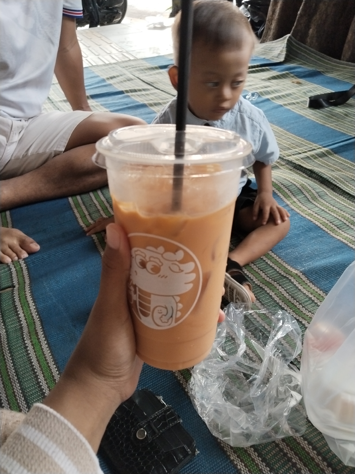 A close-up, over-the-shoulder shot shows a person's hand holding a plastic cup of iced tea. The cup has a cartoon dragon design. In the background, a young child wearing a blue shirt sits on a blue and green striped mat. A person's legs in white shorts and a white shirt are visible on the left. The scene is likely outdoors, with the mat suggesting a casual setting. The image was taken in Kecamatan Pati, Indonesia.