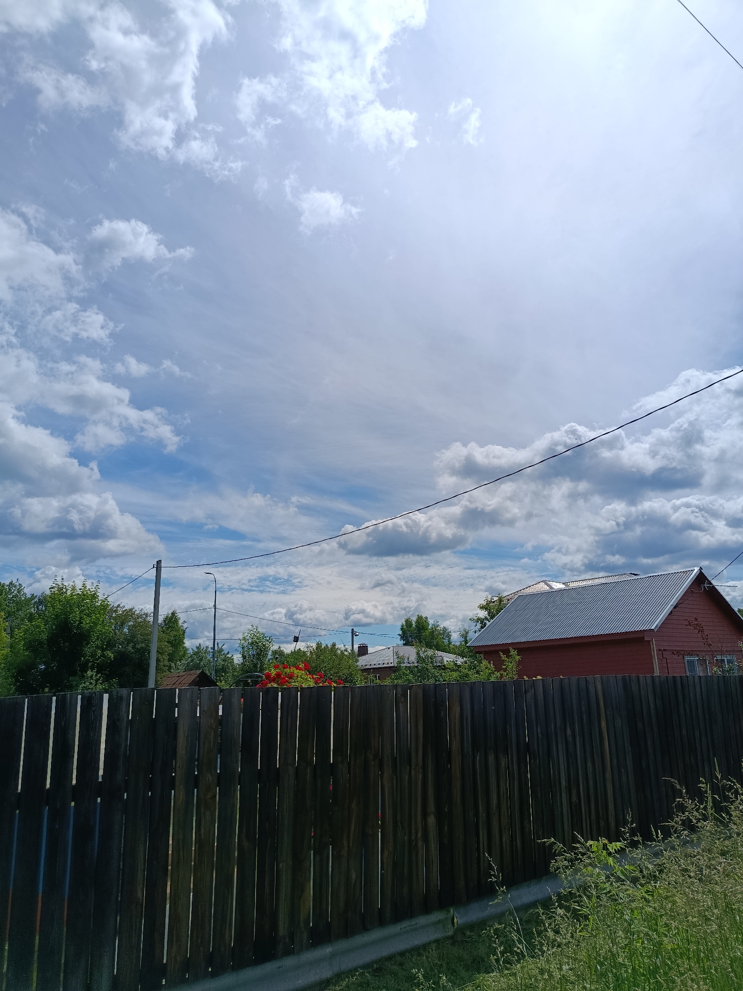 This media file depicts a rural outdoor scene in Vereya, Russia, under a bright, partly cloudy sky. A tall, dark wooden fence dominates the foreground, its vertical planks creating a textured barrier. Beyond the fence, lush green trees and foliage form a dense backdrop. Partially visible amongst the trees are rooftops and structures, including a prominent red-sided house with a corrugated metal roof. A power line stretches diagonally across the upper portion of the image, adding a man-made element to the natural landscape. The bright sky suggests it is daytime, likely late morning or early afternoon, with the sun filtering through gaps in the scattered white clouds. The overall impression is one of a tranquil, sunlit day in a village setting. No people or text are visible in the image.