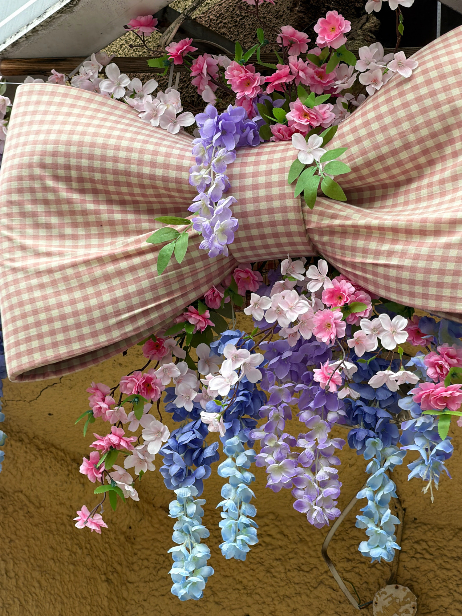 The image depicts a close-up of a large, pink and white checkered bow decorated with artificial flowers in shades of pink, white, purple, and blue. The bow is positioned outdoors, possibly as a decoration. In the background, a textured yellow wall and a slatted structure are visible. The location in the city of Koszalin, Poland is not discernible from the visual information provided. There are no people or animals present in the image, and no actions or interactions are depicted. The overall scene is a static display of decorative elements.