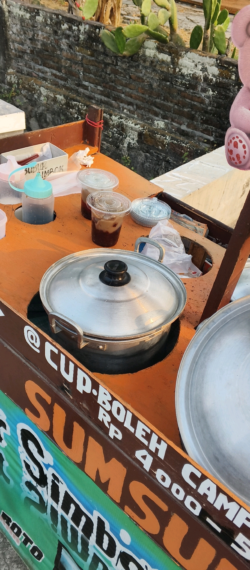This image shows a street food cart in Japanan, Indonesia. The cart is made of wood and has several compartments. On the cart, there are two plastic cups filled with a brown liquid and some solid ingredients, likely a beverage or dessert. A turquoise squeeze bottle containing a dark liquid is also visible. A large, silver pot with a black handle sits in the center of the cart, suggesting it is used for cooking or keeping food warm. In the background, there is a brick wall with some plants growing on it, including cacti. A pink teddy bear is partially visible on the right side of the image. The text on the side of the cart reads "@CUP. BOLEH SUM... RP 4.000-" and "SUMBOTO". The overall scene depicts a vendor stall set up outdoors, likely in a public area.
