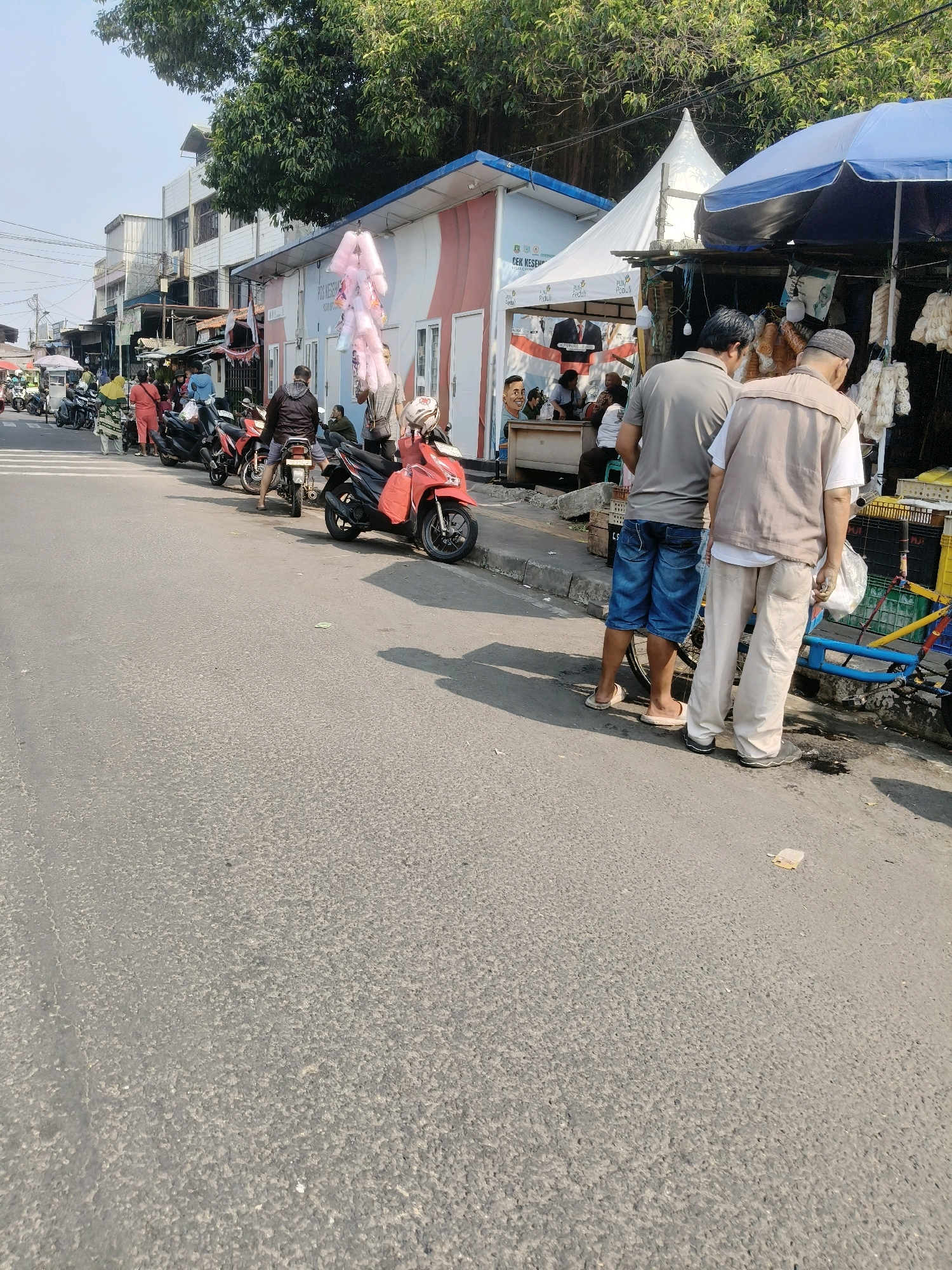 In the city of South Tangerang, Indonesia, on a sunny day, a street market scene unfolds. Several motorcycles are parked along the roadside, some with riders seated on them. People are walking and interacting with vendors. In the foreground, two men are standing, facing away from the camera. One is wearing a grey t-shirt and blue denim shorts, the other a beige vest over a white shirt and light-colored pants. Behind them, a blue and yellow cart is visible. Further down the street, a temporary stall with a blue umbrella is set up, displaying goods. To the left, a white building with red accents stands adjacent to a white tent. A string of pink cotton candy is hanging from a pole. The street is paved and shows signs of wear, with some debris scattered on the surface. Trees with dense foliage overhang the scene from above. The environment suggests a bustling urban area with commercial activity.