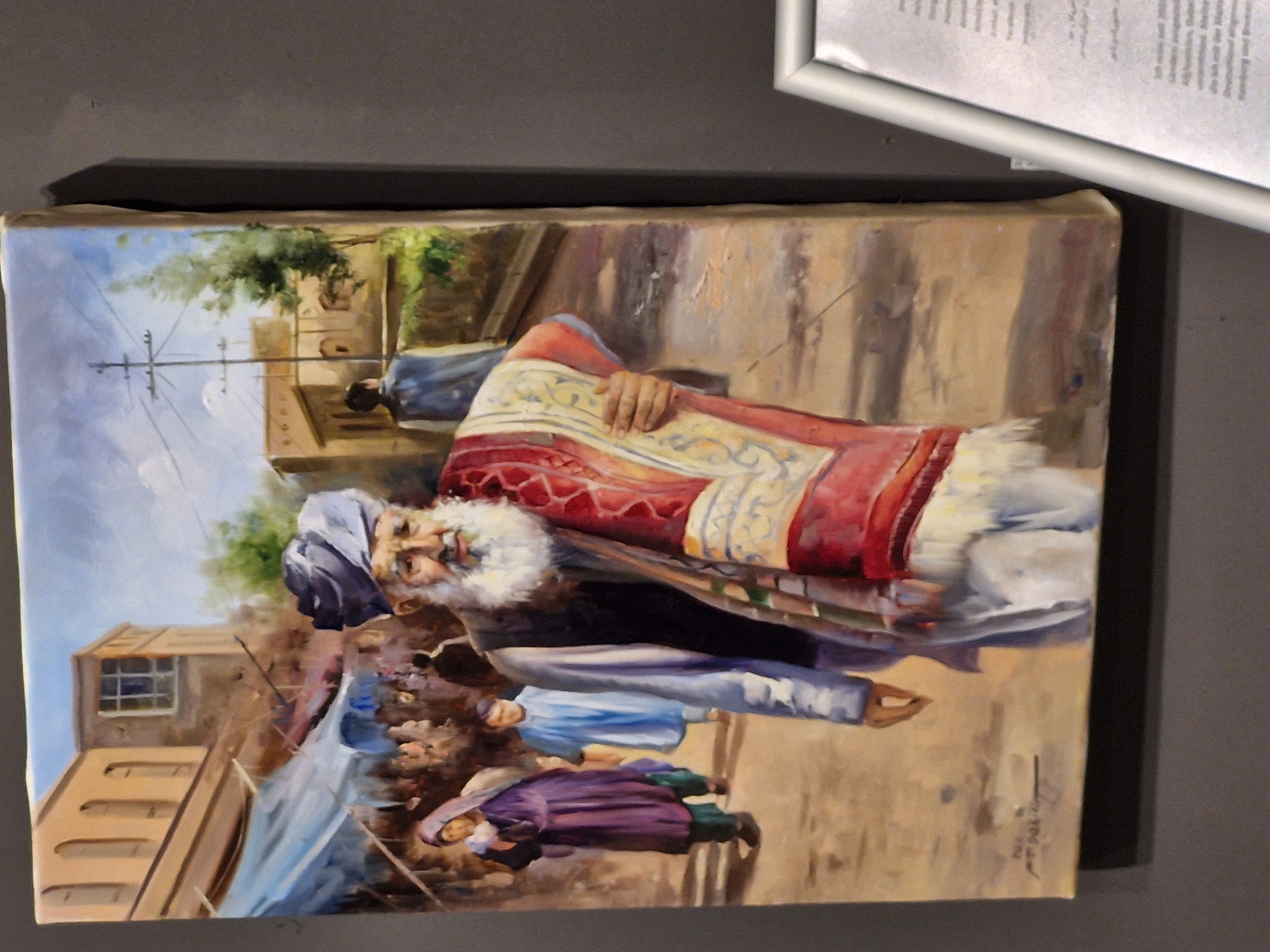 The image depicts a painting of an outdoor market scene. A bearded elderly man wearing a turban and a dark vest over a light-colored garment is walking towards the viewer, carrying a rolled-up rug with red and yellow patterns. Behind him, other figures are visible, including a woman in a purple garment and headscarf, and a boy in a blue shirt. The background shows buildings with arched windows and a sky with clouds. The painting is displayed in a museum setting, with a portion of a museum label visible in the lower right corner. The text on the label appears to be in German and mentions "Bremen." This suggests the painting is either located in Bremen or is part of a collection in Bremen that is on display.