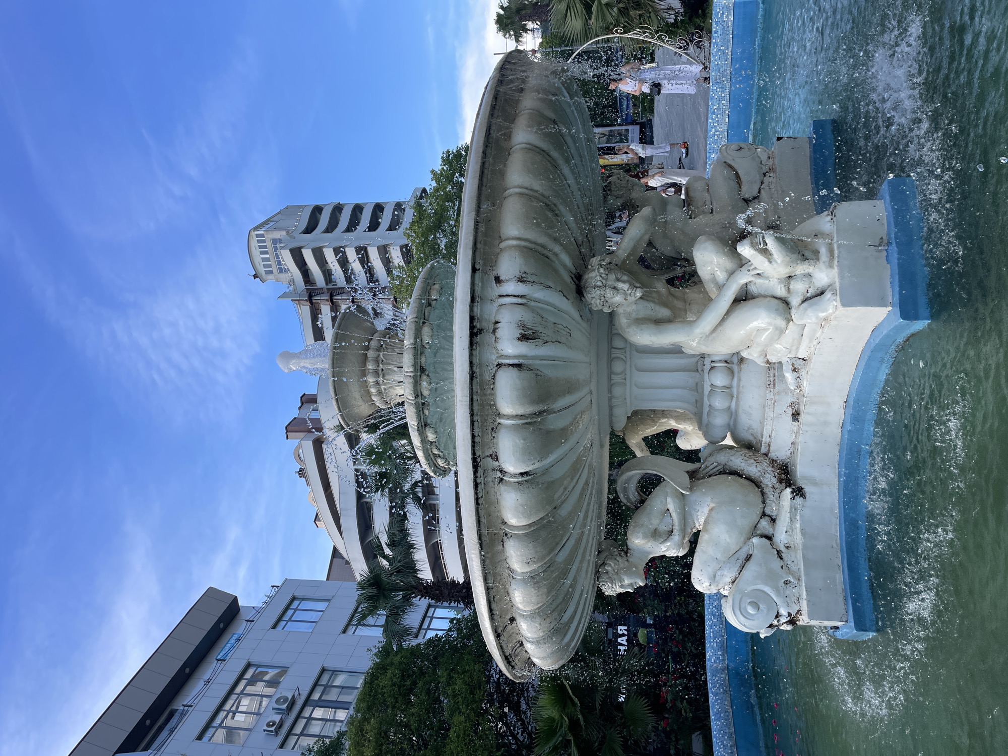 This is a medium shot, eye-level, vertical photograph of a decorative fountain in the city of Sochi, Russia. The fountain is made of white stone and features multiple tiers with sculpted figures of people and mythical creatures, some of which appear to be mermaids or mermen. Water sprays from the top tier, cascading down into the basin, which is filled with green water and illuminated by sunlight. The fountain is situated in an urban setting, with modern buildings visible in the background, including a multi-story building with curved balconies and a taller building with a distinct architectural design. Palm trees and other greenery are also present, suggesting a warm climate. Several people, dressed in casual summer clothing, are visible in the background, some standing and observing the fountain, while others are walking. The sky is clear and blue with a few wispy clouds. The overall impression is of a public space, likely a park or promenade, designed for recreation and aesthetic appeal.