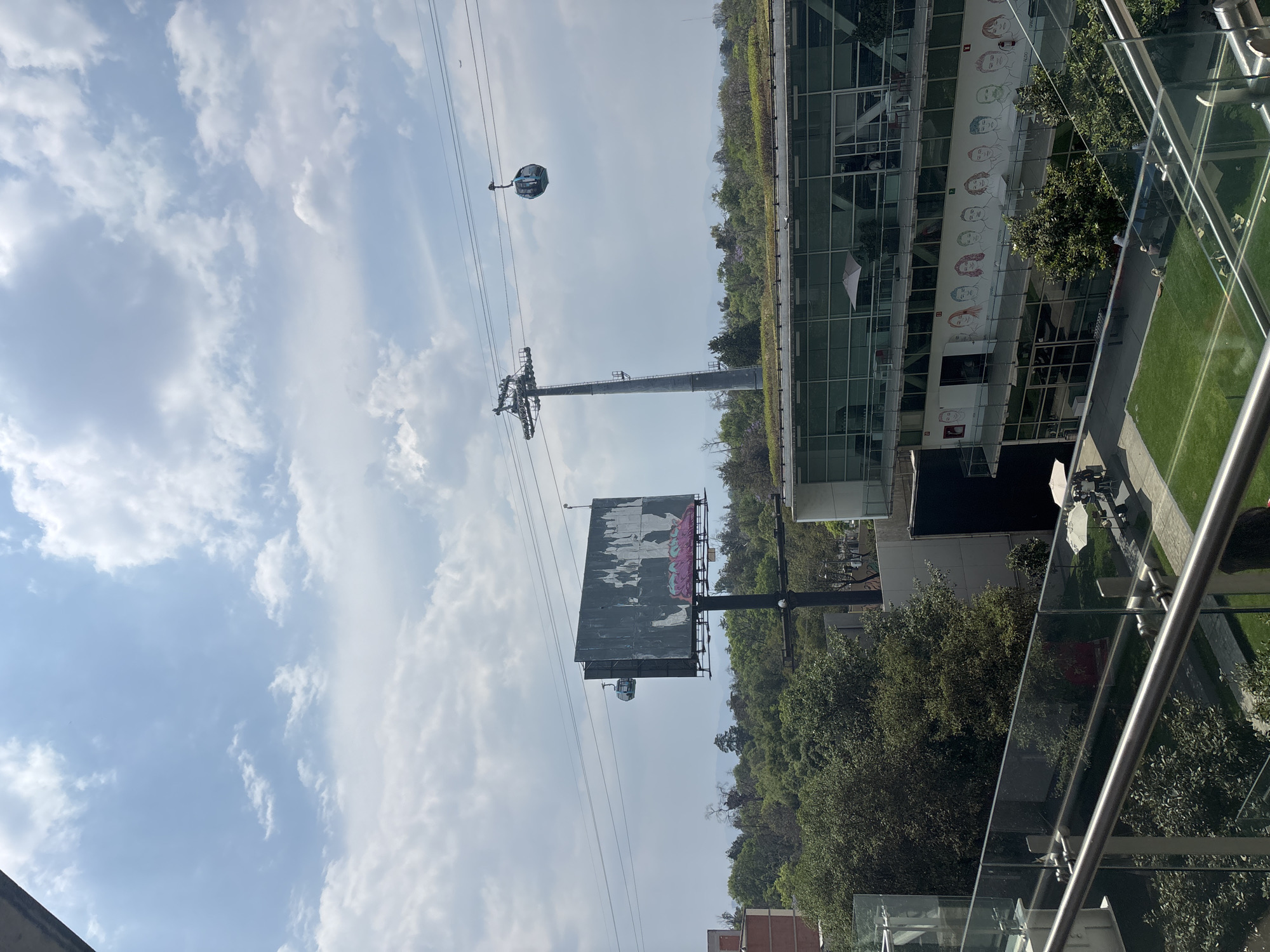 The image captures an elevated view of a modern urban landscape in Mexico City, featuring a cable car system against a partly cloudy sky. In the foreground, a sleek glass and metal railing suggests the viewpoint is from a building or balcony.
A prominent cable car pylon stands in the middle ground, supporting multiple cables along which gondolas travel. One blue and gray cable car is visible in the upper left, appearing to move along the wires, while another smaller, darker gondola is further down near a large, weathered billboard. This billboard displays faded white patches and distinct pink and possibly blue graffiti.
To the right, a multi-story modern building with extensive glass facades is seen. Its upper exterior wall features a colorful mural composed of stylized, diverse illustrated faces arranged in a row, suggesting a public art installation or branding. Below this building, lush green trees and landscaped areas are visible, with distant hills forming the horizon. The bright, clear sky with scattered white clouds indicates it is daytime, likely mid-morning or afternoon. The overall scene depicts a functional urban environment with transportation infrastructure, green spaces, and modern architecture.