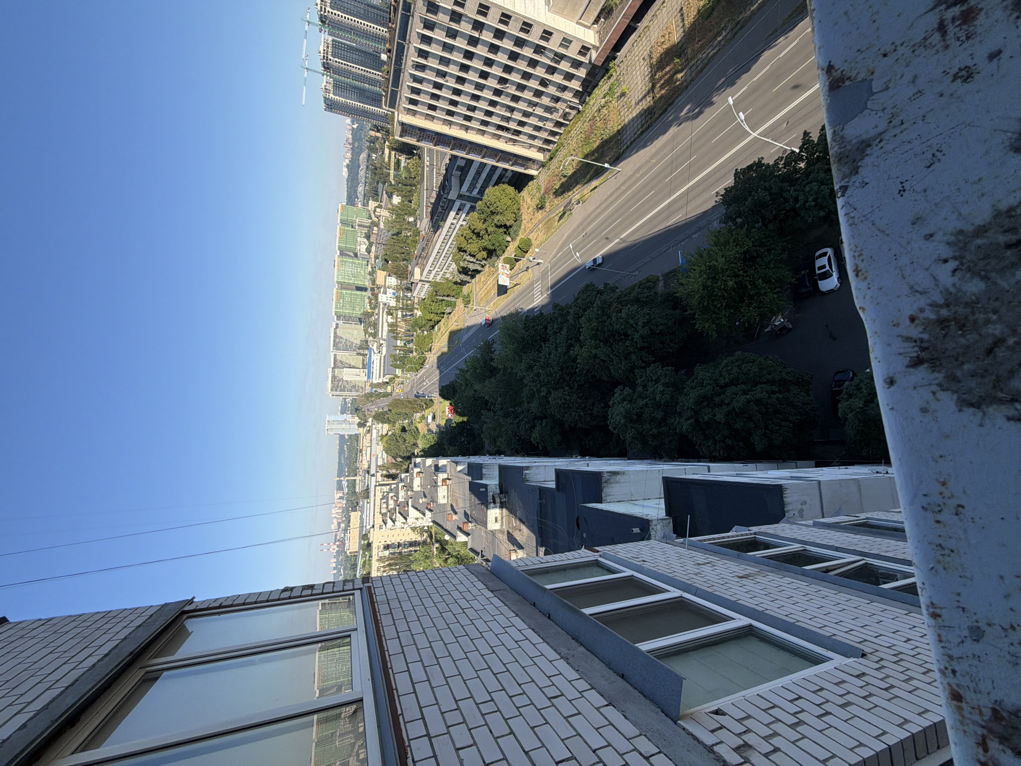 The image shows a high-angle view of a city street in Kyiv, Ukraine, likely taken from a tall residential building. The foreground is dominated by the side of a white brick building with several windows. To the right of this building, a multi-lane road stretches into the distance, lined with trees and various buildings. In the background, a cluster of tall, modern apartment buildings with green facades is visible, along with other urban structures and construction cranes. The sky is clear and blue, indicating a sunny day. Several cars are parked along the side of the road and one is driving on the street. There are no people visible in the image. The scene depicts a typical urban environment with a mix of residential and commercial architecture.