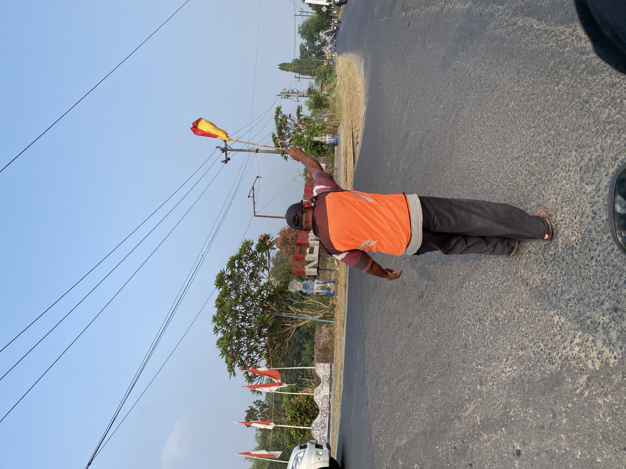 A person wearing an orange vest and a black baseball cap is standing on an asphalt road, facing away from the camera, and holding a red and yellow flag on a pole. The person's left arm is raised, holding the flag. The road is cracked and appears to be a two-lane street with no visible lane markings. In the background, on the left side of the road, there are several Indonesian flags (red and white) on poles, a white car, and a decorative wall with a mosaic pattern. Further back, there is a lush green tree and some buildings. On the right side of the road, there are power lines, utility poles, and more trees. The sky is clear and blue. The scene appears to be set in a rural or semi-urban area, possibly on the outskirts of Kasihan, Indonesia, given the presence of Indonesian flags and the overall landscape.
