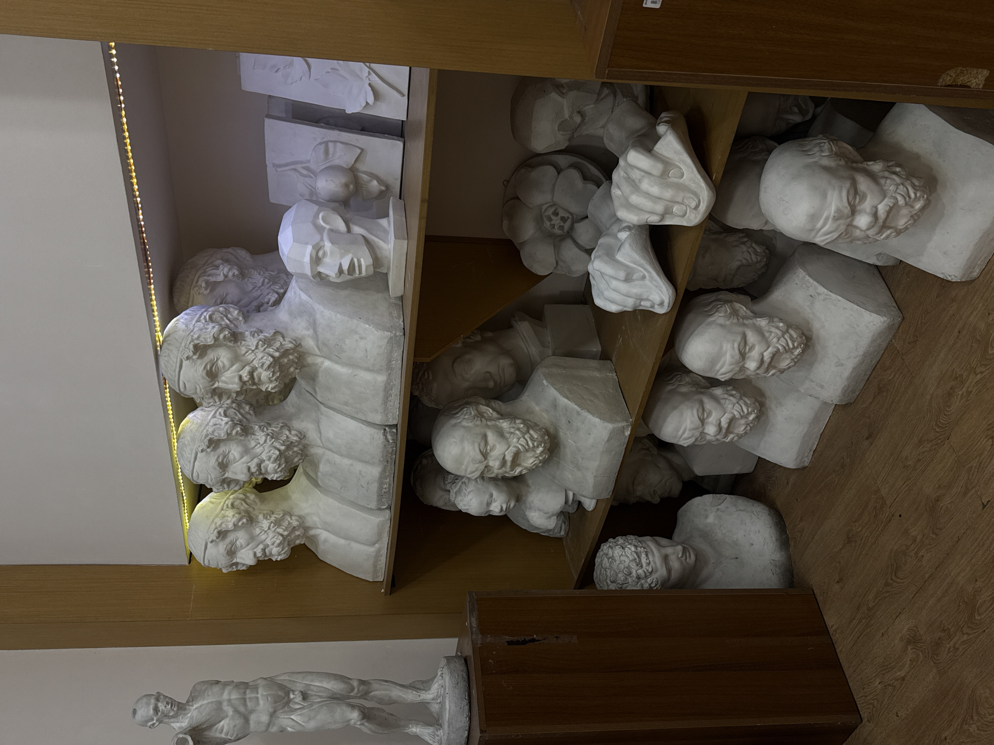 The image depicts a collection of plaster casts of classical sculptures stored on wooden shelves. The setting appears to be a storeroom or studio, potentially within a museum or educational institution. The sculptures consist mainly of busts and heads, with one full figure on the bottom left. The lighting is mixed, with some artificial illumination along the edge of the shelf. The exact location in Yerevan, Armenia is not discernable from the image.