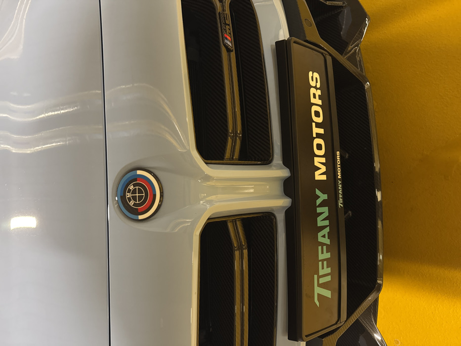The image displays a close-up, head-on view of the front fascia of a light-colored (possibly light grey or silver) BMW M2 vehicle. The prominent BMW emblem on the hood features the M 50th anniversary design with concentric semicircles in light blue, dark blue, and red around the traditional BMW logo. Below this, the vehicle's double kidney grille is visible, filled with a black carbon fiber textured mesh and accented by horizontal golden-bronze-colored slats. A small "M2" badge is affixed to the upper right section of the grille. Directly beneath the grille, a black license plate holder is installed, bearing the inscription "TIFFANY MOTORS" in light green/teal and gold lettering. The words "TIFFANY MOTORS" are repeated in smaller text below the primary inscription. Further down, the lower bumper features additional exposed carbon fiber elements. The vehicle appears to be in an indoor environment, evidenced by reflections on its polished surfaces and a distinct, solid yellow-orange floor visible at the bottom of the frame. Based on the "Tiffany Motors" branding, the scene is situated at a Tiffany Motors luxury car dealership in Warsaw, Poland. No individuals are present in the image, and no actions or interactions are depicted.
