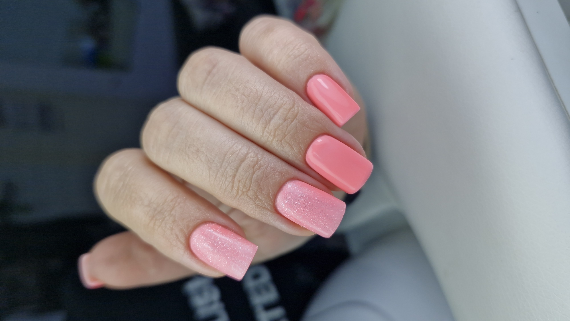 A human hand is positioned in the center of the frame. The hand is light-skinned with short, manicured nails painted in two different shades of pink. Two nails are a solid, vibrant pink, while the remaining three have a lighter, shimmery pink finish. The fingers are slightly curled, creating some wrinkles in the skin. The background is blurry and indistinct, but appears to include a dark object and part of a white surface. The scene does not provide any information about the location of the city of Otchor, Russia.