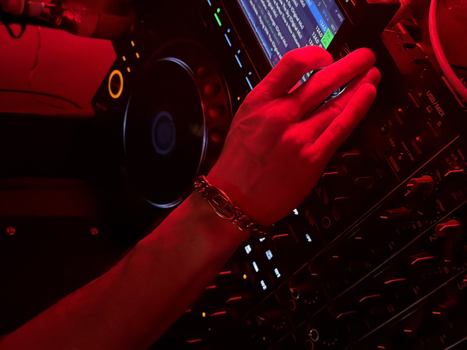 The specific location within Yerevan, Armenia, is not discernible from this image.
The scene depicts a close-up, high-angle view of an individual's right arm and hand interacting with professional DJ equipment, bathed in intense red ambient light. The individual wears a dark, buttoned shirt and a metallic, chunky chain-link bracelet on the right wrist, featuring an oval-shaped decorative link. The right hand, with fingers slightly curled, is positioned over the controls of a multi-channel DJ mixer, suggesting active operation. The mixer displays numerous faders, rotary knobs, and backlit buttons. A digital screen on the mixer shows track information, including titles such as "Paying With You (Original Mix)" and tempo, indicated as "124.0 BPM". To the right of the arm, a portion of a CDJ or media player is visible, featuring a large circular jog wheel with a blue illuminated center and an orange illuminated outer ring. In the upper right background, a clear glass bottle containing a reddish liquid is partially visible. The overall environment is dark, characteristic of a nightclub or bar setting.