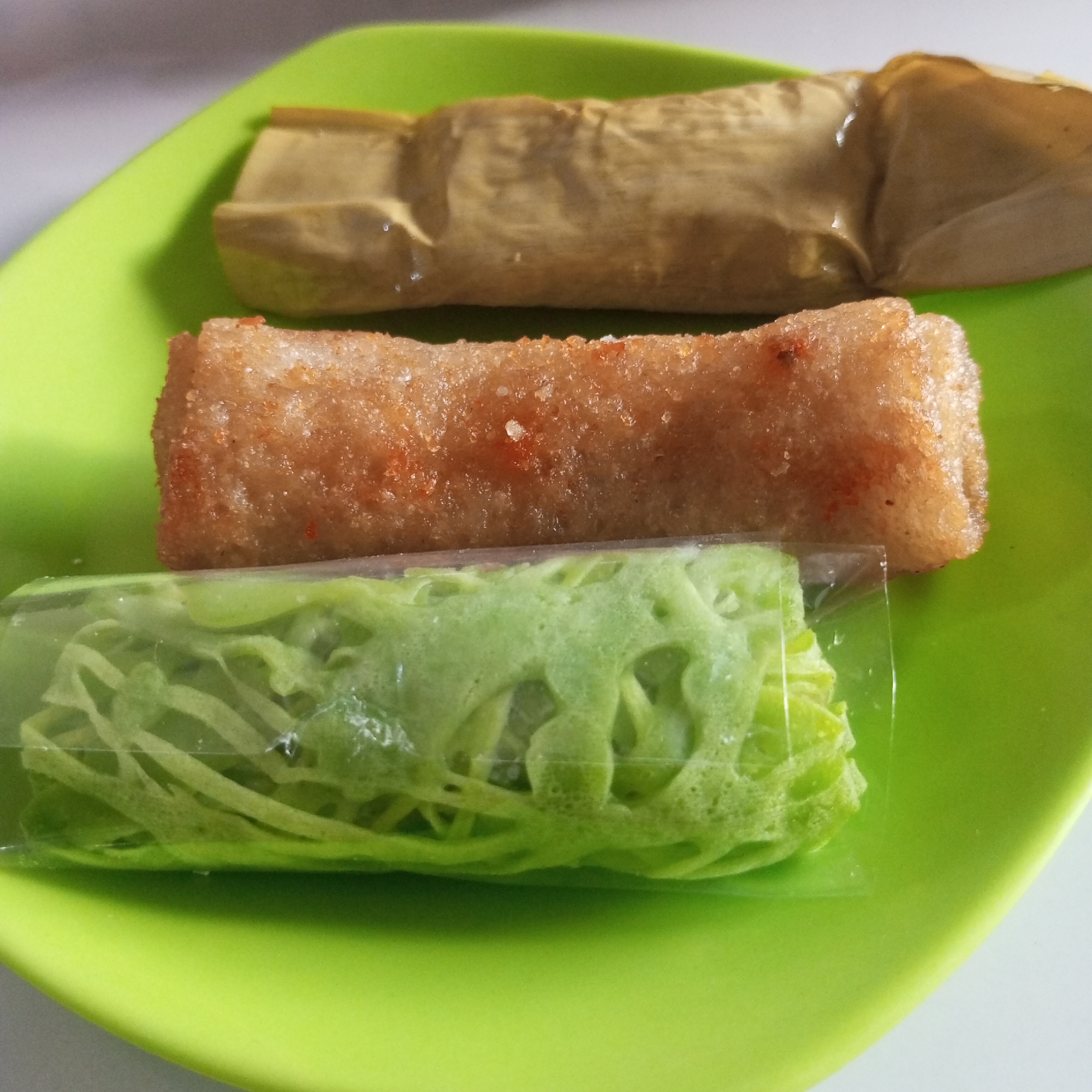 This image displays three distinct food items arranged horizontally on a bright lime-green plate. The scene is static, likely indoors, with a soft, neutral-toned background that is out of focus.
The topmost item is a cylindrical food product encased in light brown, creased wrapping, possibly a dried leaf or parchment paper. Its texture appears somewhat soft beneath the wrapper, and there are subtle reflections indicating a slight sheen.
Beneath it, the middle item is a golden-brown, elongated, fried food piece. Its surface is notably textured, suggesting a crispy exterior, possibly coated with fine granular elements that give it a speckled appearance with hints of reddish-orange.
The bottommost item is a vibrant green, intricate, web-like or lacy pastry, enclosed within a clear plastic wrapper. The wrapper shows some reflections and slight wrinkling, but the delicate, porous structure of the green food is clearly visible through it.
All three items are positioned parallel to one another on the green plate, which has a slightly raised edge. The lighting is even and bright. This scene is located in Lhokseumawe, Indonesia.