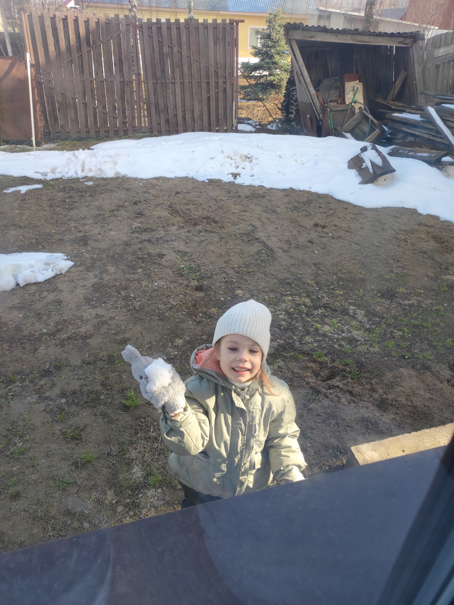 A young girl, possibly between 4-6 years old, stands outdoors in what appears to be a backyard or rural area, possibly in Votkinsk, Russia. Patches of snow are visible on the ground, suggesting it is either late winter or early spring. The girl is wearing a white knit hat, a light green winter jacket, and gloves. She is holding a stuffed toy in her gloved hand with a clump of snow on it. The girl is smiling slightly. In the background, a wooden fence and a shed-like structure are visible. Some wood and discarded materials are stacked by the shed. The ground is mostly dirt and sparse grass. The perspective is from slightly above. The bottom right corner is dark, perhaps the edge of a window frame.