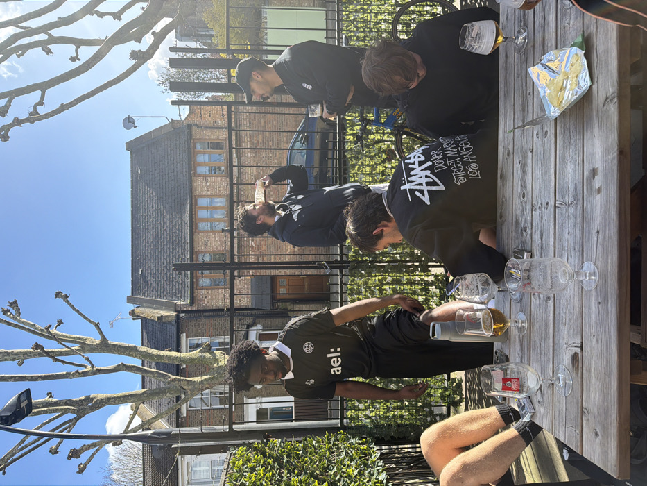 The scene is an outdoor seating area, likely a pub garden or patio, on a clear, sunny day in London, United Kingdom, evidenced by the characteristic terraced brick houses and pollarded trees in the background. Six adult males are present.
In the foreground, a wooden slatted picnic table holds several stemmed beer glasses, some containing an amber liquid and others empty. Also on the table are a cylindrical grey water bottle, a small rectangular white box (possibly cigarettes or a lighter), a dark-colored smartphone, and an open foil packet of plain potato crisps.
Three males are seated at the table. On the far left, only the lower legs of a person in shorts, black socks, and black trainers are visible. In the middle, a male with dark hair, facing away, wears a black sweatshirt featuring "Stüssy DONER STREET MARKET LOS ANGELES" on the back, and is looking downwards. On the right, a male in a dark long-sleeved top also looks downwards, with a glass of amber liquid in front of him.
Three males are standing. On the left, a male with dark curly hair wears a black short-sleeved jersey with a white collar, a white logo, and "ael:" printed in white. He stands with his left foot propped on his right knee, looking downwards. In the middle-right, a bearded male in a dark hooded top is drinking from a tall glass containing an amber liquid. To his right, a male wearing a black baseball cap and a dark top with a white logo holds a glass of amber liquid and faces towards the bearded male.
The background features a black wrought-iron fence. Beyond the fence, a row of two-story terraced brick houses, common in London, are visible. Bare-branched, pollarded trees stand prominently, alongside a street lamp. A partially visible blue bicycle is present behind the seated men, and a dark car is visible behind the fence. The sky is clear blue with minimal cloud cover.