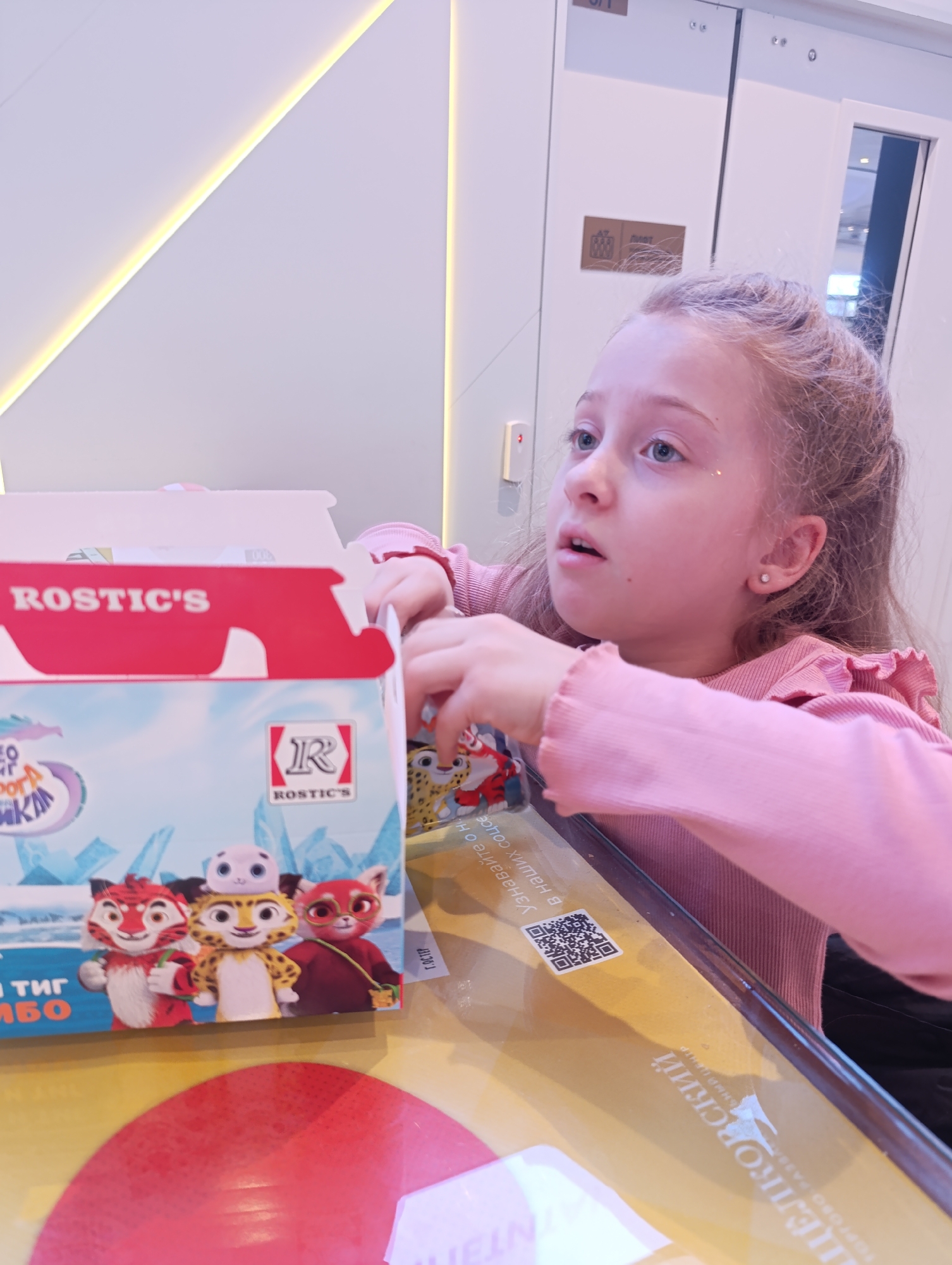 The image depicts a young girl with fair hair and blue eyes sitting at a table in a location identified as "TSELKOVSKIY Shopping and Entertainment Center" in Moscow, Russia. She is wearing a pink ruffled long-sleeved shirt. In front of her is an open box from "Rostic's", a fast-food restaurant, with cartoon characters printed on the side. There's a table with a reflective surface, and a QR code is visible on a label on the table. In the background, there is a modern interior with a wall featuring a geometric design and a doorway.