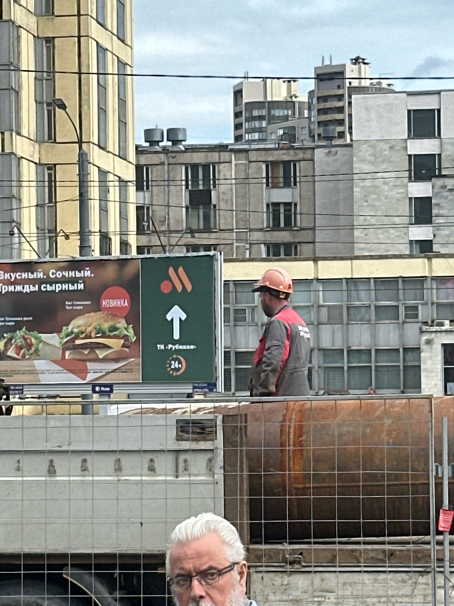 In Murino, Russia, a construction worker wearing a hard hat and a red and grey jumpsuit stands near a billboard. The billboard features a large image of a burger and fries, with Russian text advertising a "new item" and "tasty, juicy, cheesy burgers." Behind the billboard, several multi-story buildings are visible, characteristic of urban Soviet-era architecture, with many windows and balconies. A street lamp is positioned on the left side of the frame, and power lines crisscross the sky. The foreground shows a section of a construction site with a rusty cylindrical object covered by a wire mesh fence, and the face of an elderly man with white hair and glasses is partially visible at the bottom of the frame.