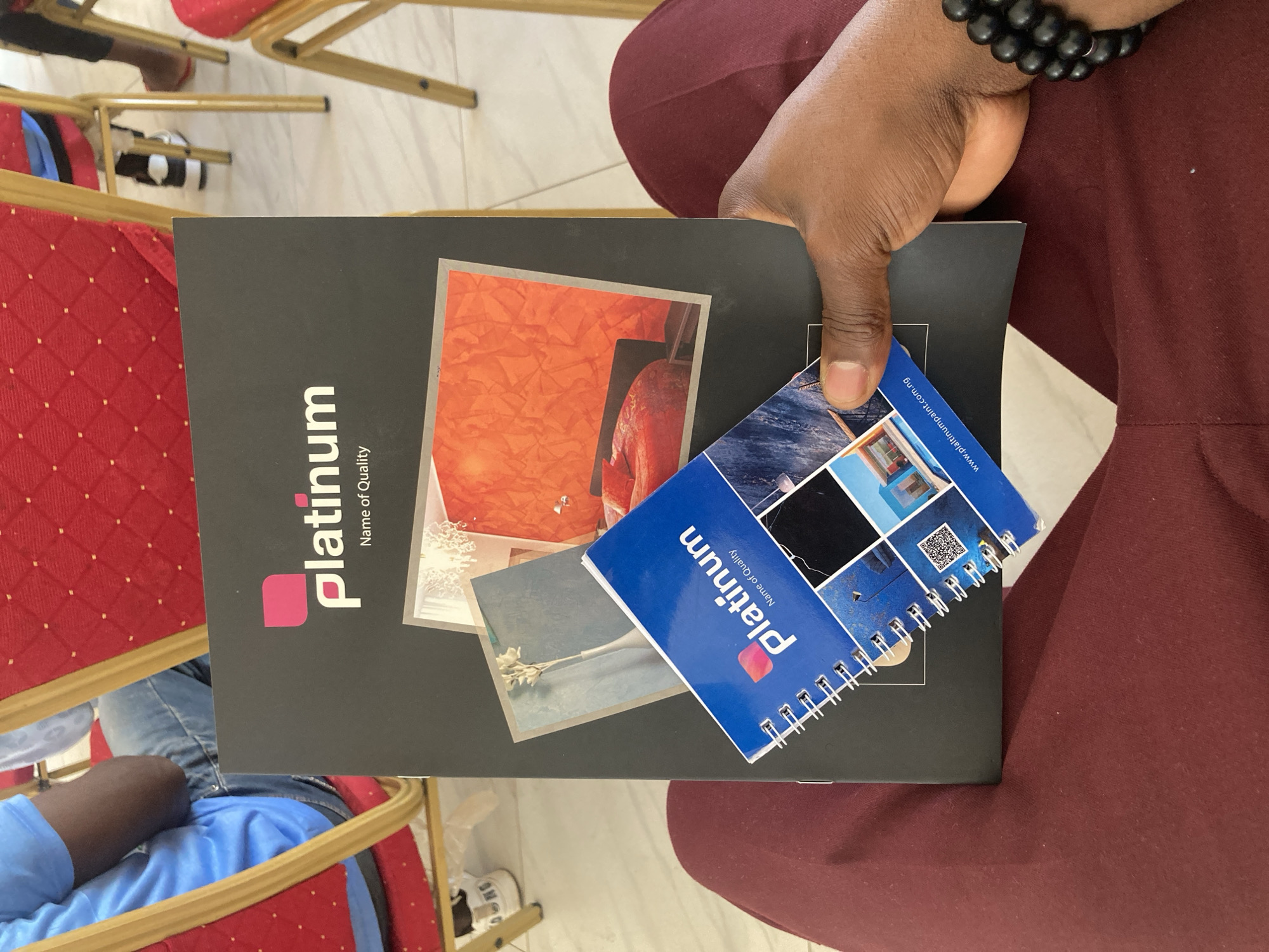 A person wearing a maroon garment and a black beaded bracelet is holding a black, rectangular Platinum brand catalog, on which are displayed three smaller, rectangular images, one containing an orange abstract design, one containing a vase of white flowers, and the third partially obscured, over a smaller blue Platinum brand notebook, which features a photograph collage. The location is Jos, Nigeria. Additional elements include chairs, clothing and a white tile floor.