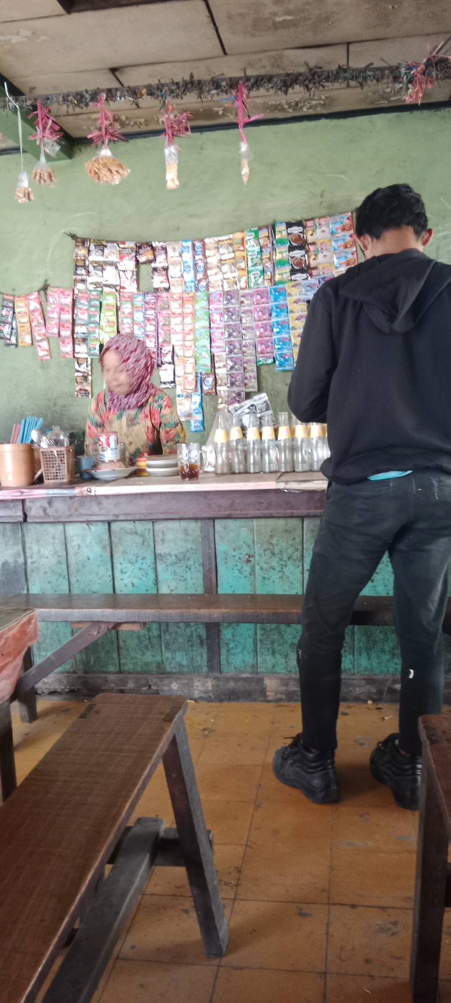 The image depicts an outdoor stall in Ngargosari, Indonesia, likely a small convenience store or warung. A woman, wearing a patterned hijab and a colorful shirt, stands behind a counter, appearing to serve a customer. The counter displays various packaged snacks and drinks. A young man, dressed in a black hoodie and dark pants, stands facing the counter, presumably the customer. The stall is simple, with wooden shelves and benches, suggesting a rustic and local setting. The overall atmosphere is casual and everyday.