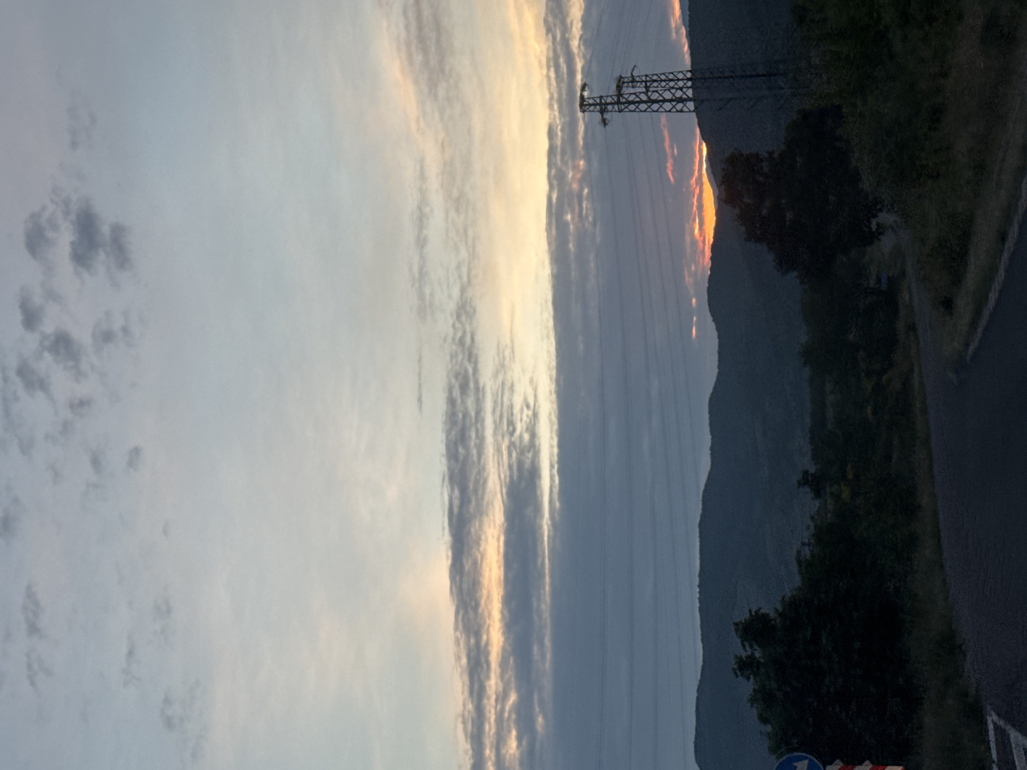 The image captures a landscape scene in Binkos, Bulgaria, at either sunrise or sunset, characterized by a dramatic sky and silhouetted terrain. The sky dominates the upper and central portions, featuring elongated clouds illuminated by warm orange and yellow light near the horizon, fading into lighter grey and pale blue further up. A striking band of bright orange light is visible peeking over the top of dark, silhouetted mountains or large hills in the mid-ground on the right.
A prominent electricity pylon stands on the right side of the image, its dark, latticed structure outlined against the sky, with multiple power lines extending horizontally across the mid-ground. Below the mountains, dense, dark green trees and vegetation line what appears to be a road or path running along the right side of the frame. In the bottom right corner, a partial view of a blue circular road sign and red and white striped barriers or cones indicates the presence of a road or construction area. There are no people visible, and no specific activities are taking place; the scene is a static depiction of a natural landscape infused with elements of human infrastructure. No legible text is visible.