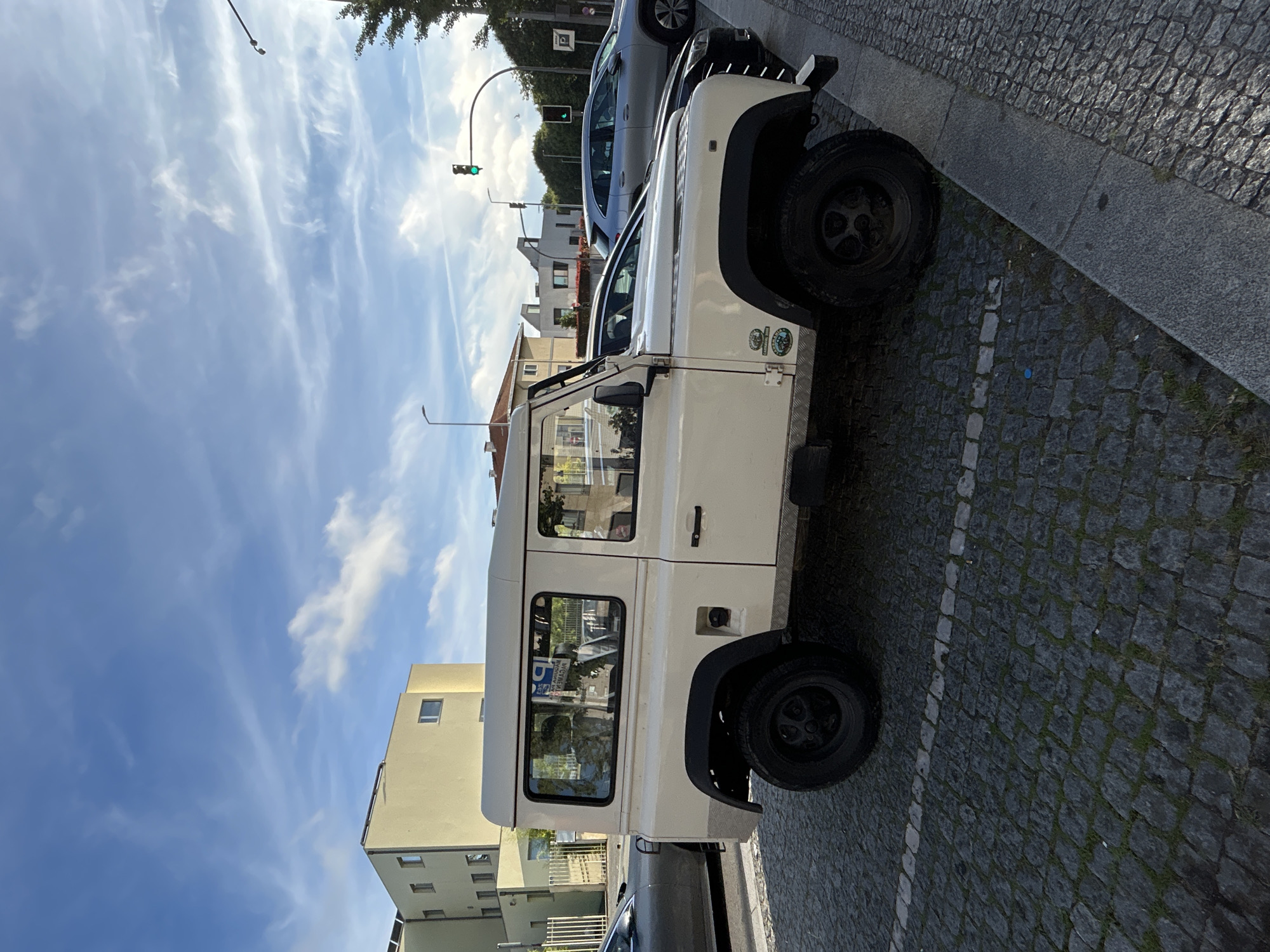 A beige Land Rover Defender is parked on a cobblestone street in Porto, Portugal. The vehicle is facing right, and its rear is towards the viewer. The Defender has a classic boxy design with large tires. Behind the Defender, several other vehicles are parked. In the background, there are buildings, traffic lights, and streetlights. The sky is blue with wispy clouds. The image appears to have been taken during daylight hours.