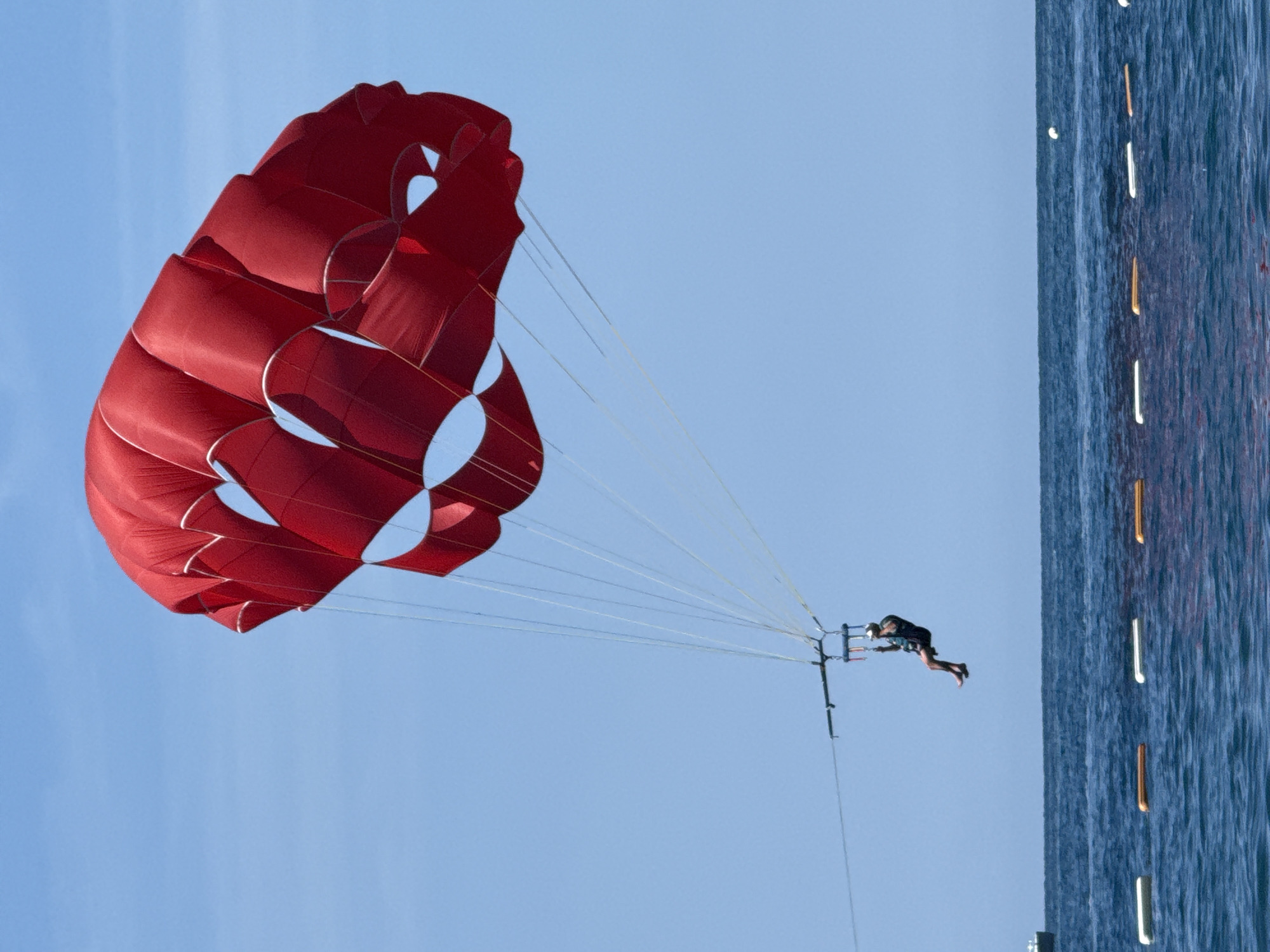An outdoor, mid-day photograph, taken in Boreti, Montenegro, captures a single individual parasailing over the Adriatic Sea. A large, bright red parasail, featuring a complex structure of inflated air cells with prominent white seam lines and scalloped edges, is prominently displayed in the upper-right quadrant of the image against a clear, light blue sky. Numerous white suspension lines descend from the canopy, converging at a black horizontal bar.
Suspended beneath this bar is a single person, seen from the torso down, seated within a black harness. The individual wears dark clothing, possibly a wetsuit, and a light-colored helmet. Their bare feet dangle, and their arms are bent at the elbows, with hands positioned near the harness straps.
Below the parasailer, the dark blue sea exhibits a gently rippled surface with subtle whitecaps. A linear arrangement of approximately six rectangular buoys, alternating between white and orange-brown, extends across the mid-ground of the water. On the far left, a small portion of a dark-hulled boat is visible near the horizon. A small, white, spherical object floats on the water's surface on the far right. The horizon line separating the sky from the sea is clear and extends horizontally across the frame.
