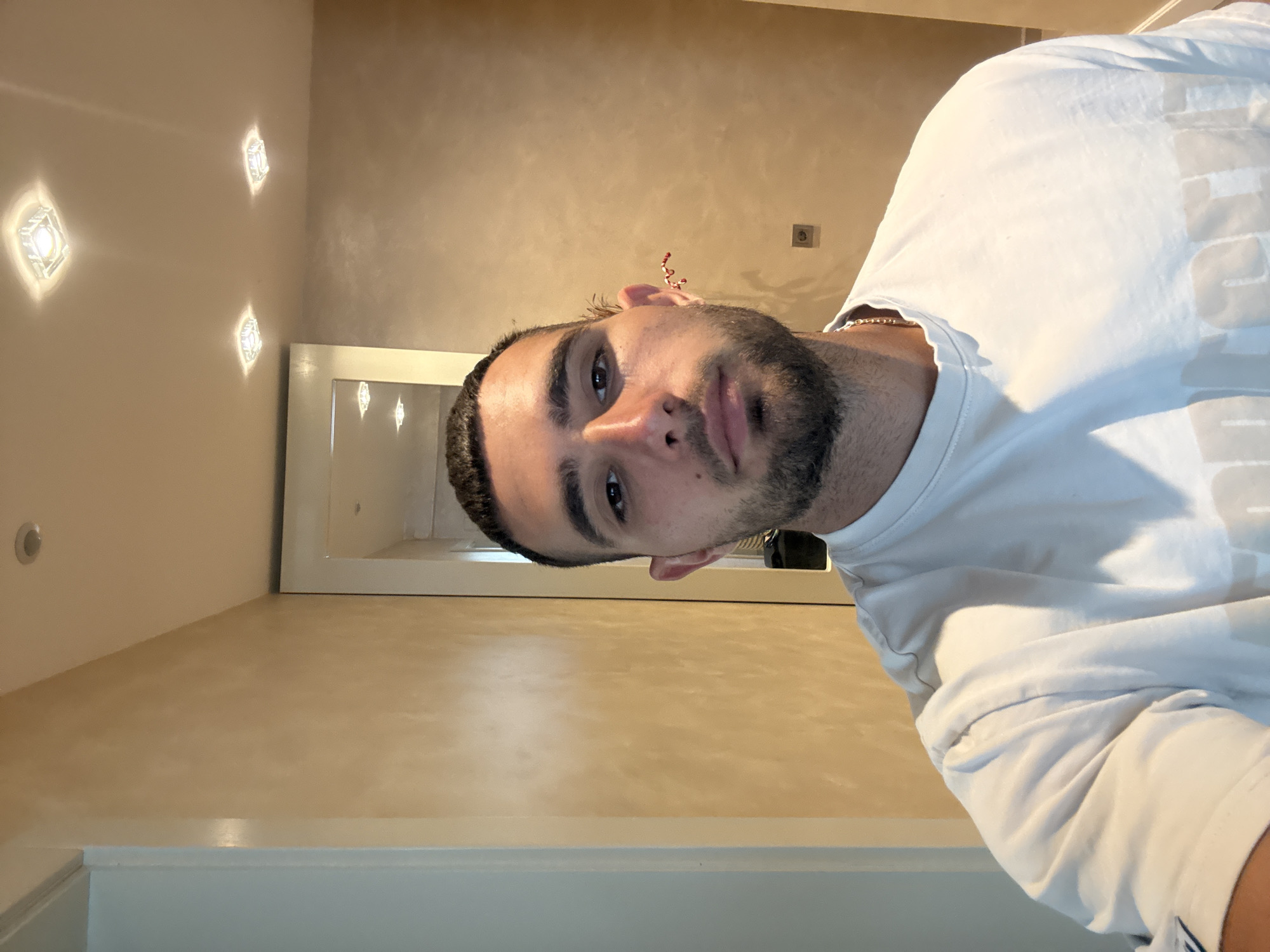 A man with dark hair and a beard is taking a selfie in an indoor hallway. He is wearing a white t-shirt and a gold chain necklace. The hallway walls are a warm, textured beige, and the ceiling has recessed square lights. There is a mirror visible in the background. The image does not provide any specific information that would allow it to be located in Gabrovo, Bulgaria.