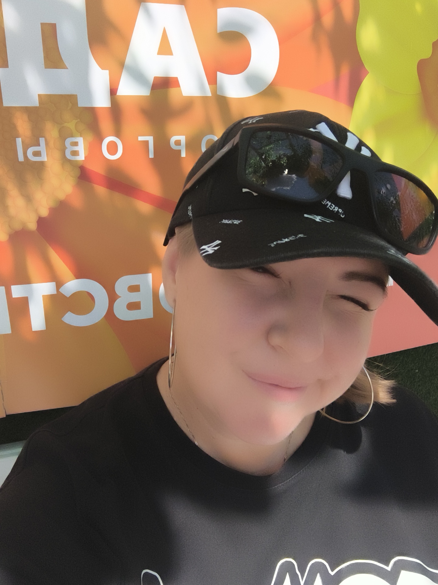 In an outdoor setting, a young woman with blonde hair is captured in a selfie. She is wearing a black t-shirt and a black baseball cap with white patterns and sunglasses perched on the brim. Large, white letters are visible in the background, part of a sign that appears to be illuminated by sunlight, casting shadows. The woman is looking at the camera and smiling, with one eye squinted. She is wearing large hoop earrings. The background suggests an urban environment, possibly a city street or park.