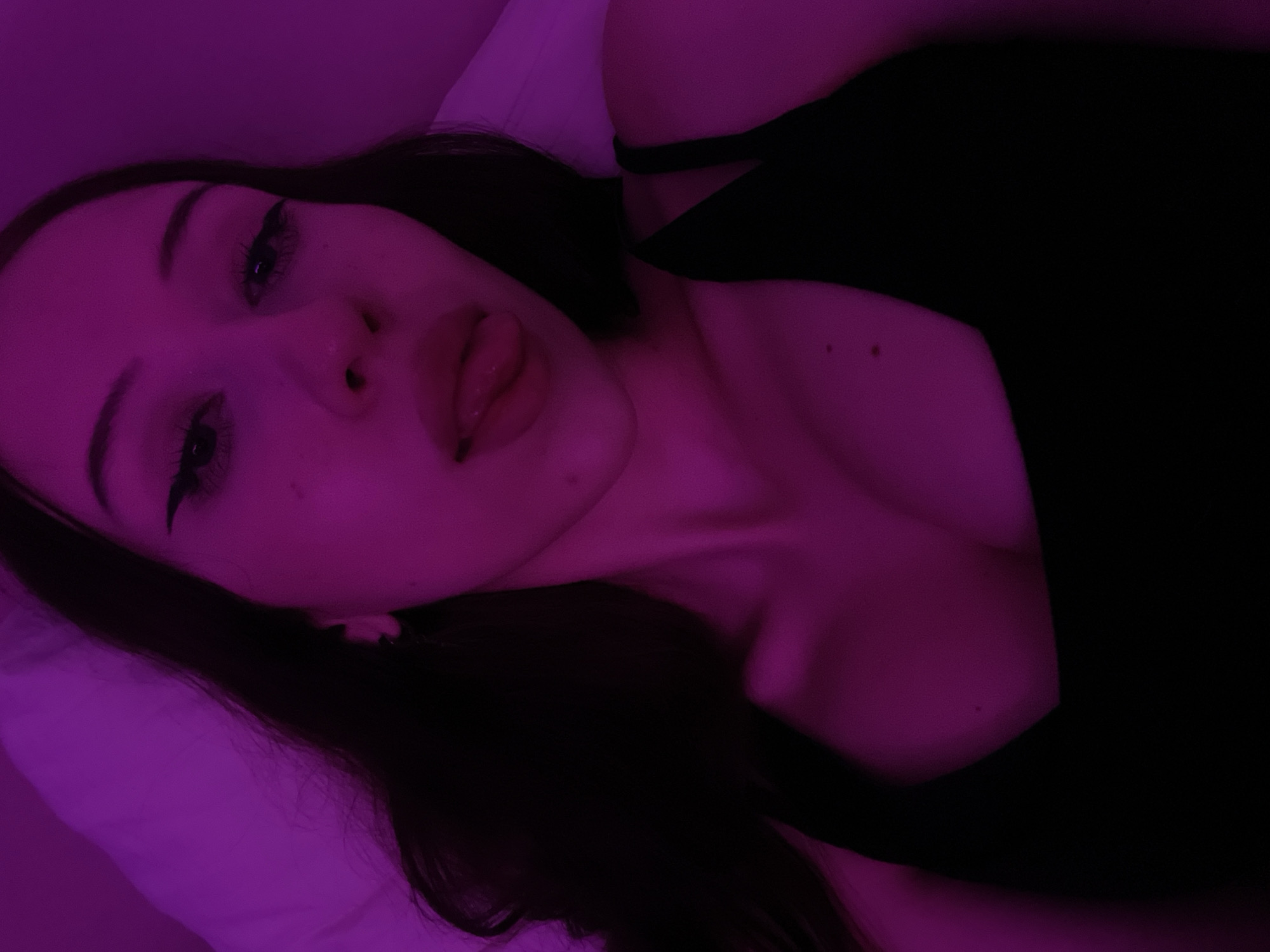 A young woman with long, dark hair and dark eyes is shown in a close-up, selfie-style shot. She is lying down, with her head tilted slightly back and her tongue extended. She is wearing a black tank top. The lighting is a strong, saturated pink or purple hue, casting a dramatic glow on her skin and hair. The background appears to be a soft, possibly white, surface, such as a pillow or duvet. There is no indication of the location within Votkinsk, Russia, or any specific environmental details that could pinpoint a location within the city. The image is a portrait focusing on the woman's face and upper body.
